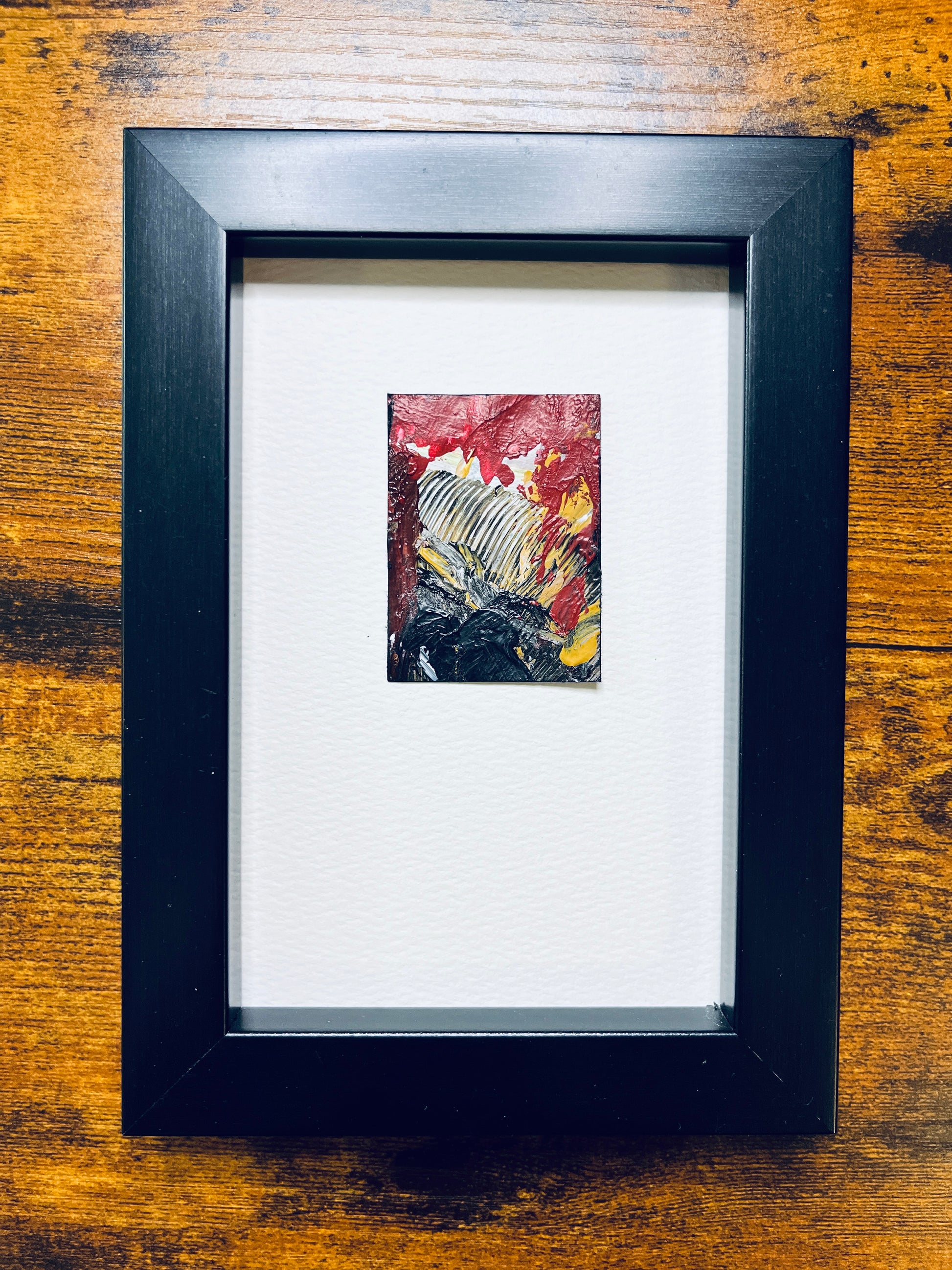 Original artwork by Anet Skillin. This miniature abstract painting is painted on Kraft paper, in black, white, red, and yellow ochre colors; mounted on 4x6-inch paper and photographed in a black 4x6-inch frame against a wooden table.