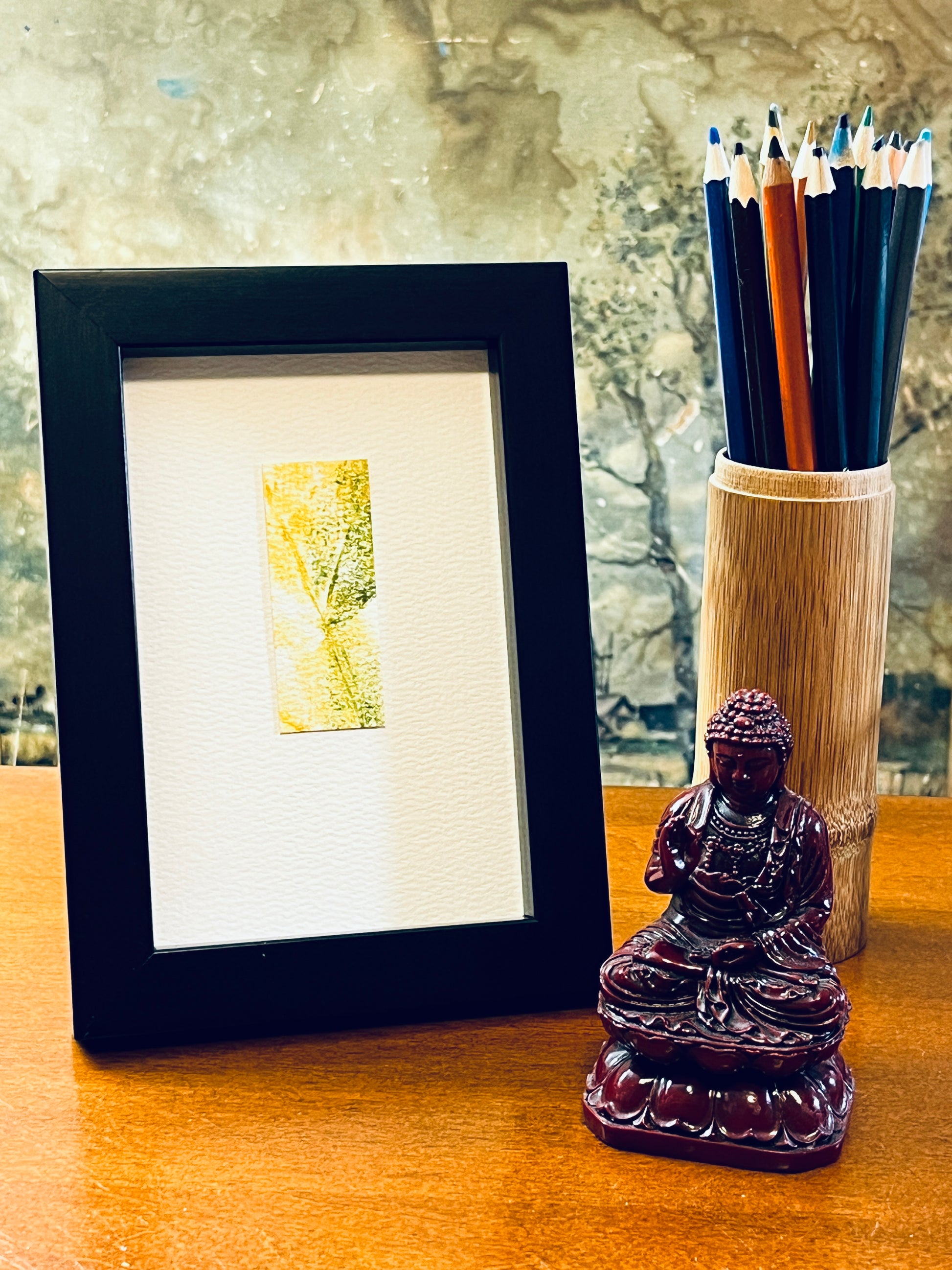 Original yellow and green watercolor leaf impression on textured cold press watercolor paper, mounted on 4x6-inch paper in portrait orientation and displayed in a 4x6-inch black frame next to a bamboo pencil holder and a small Buddha statue. 