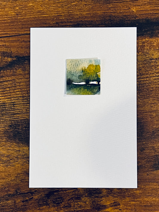 Original square miniature watercolor painting of an abstract landscape with a maple or oak tree in yellow ochre, green, and sepia foliage beneath cloudy blue skies. Painted on textured cold press watercolor paper, mounted on 4x6-inch paper in portrait orientation, and photographed against a wooden table.