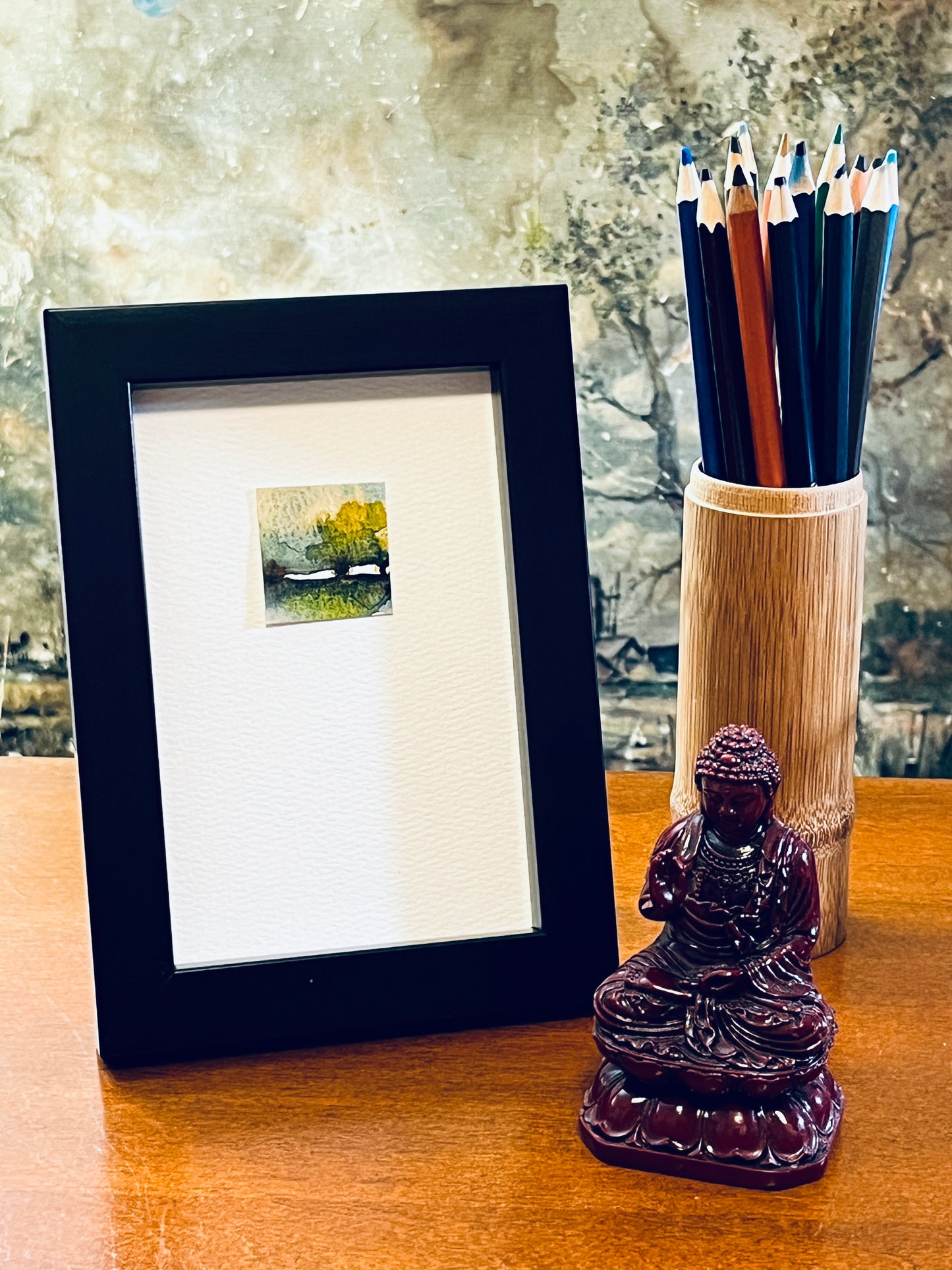 Original miniature watercolor painting of an abstract landscape with a maple or oak tree in yellow ochre, green, and sepia foliage beneath cloudy blue skies. Painted on textured cold press watercolor paper, mounted on 4x6-inch paper in portrait orientation. Displayed in a black 4x6-inch frame next to a bamboo pencil holder and a small Buddha statue.