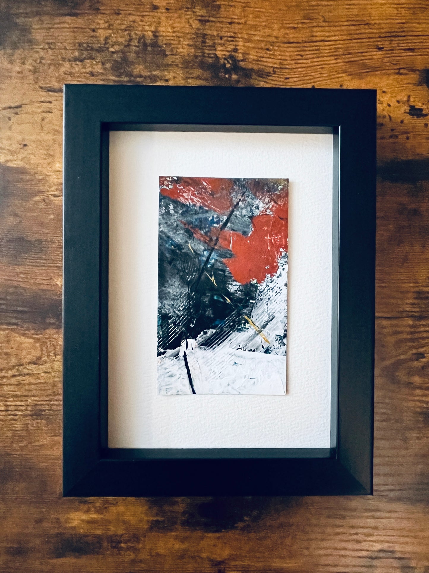 Original artwork by Anet Skillin. This miniature abstract is painted on recycled packaging in white, red, green, blue and yellow-ochre colors. Photographed matted in a black 5x7-inch frame against a wooden table.