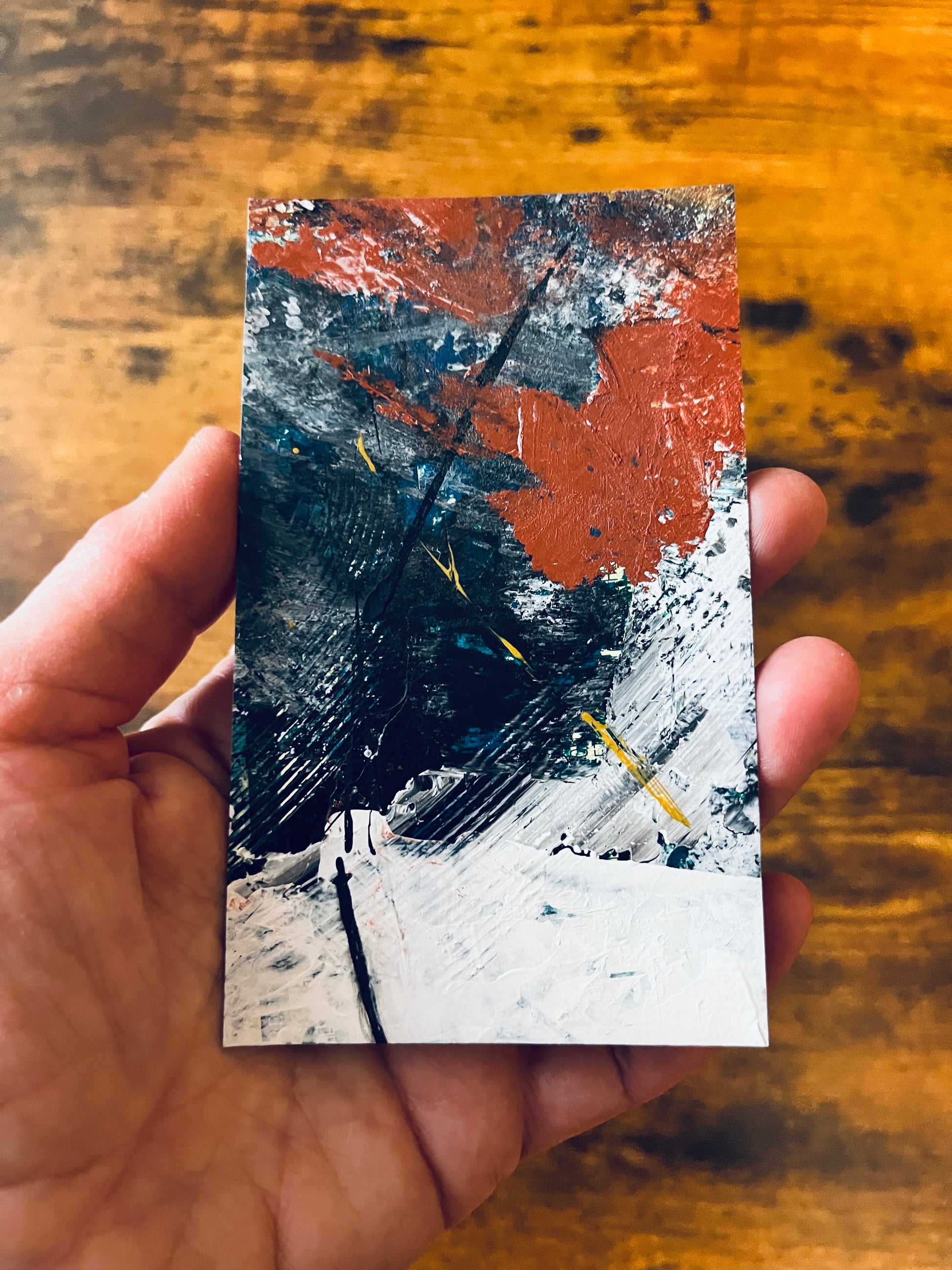 Original artwork by Anet Skillin. This miniature abstract is painted on recycled packaging in white, red, blue and yellow-ochre colors. Photographed in the artist's hand against a wooden table.