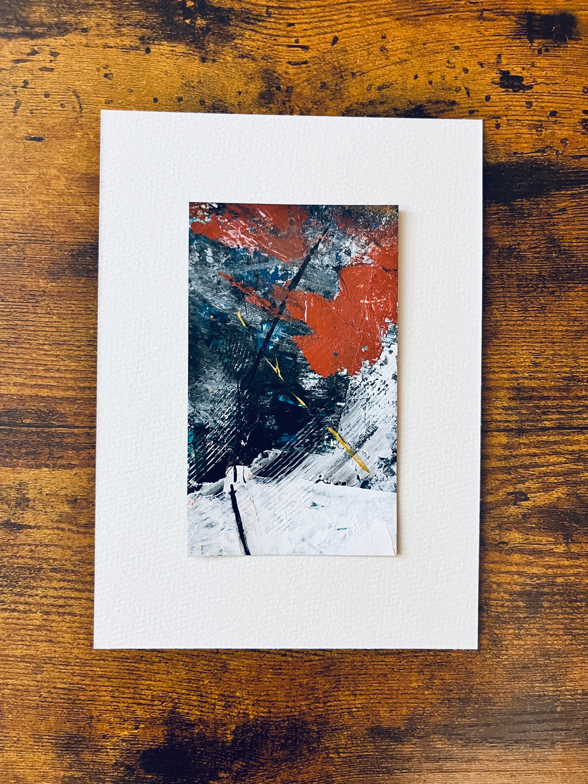 Original artwork by Anet Skillin. This miniature abstract is painted on recycled packaging in white, red, blue and yellow-ochre colors; displayed on a 5x7-inch sheet of paper and photographed against a wooden table.