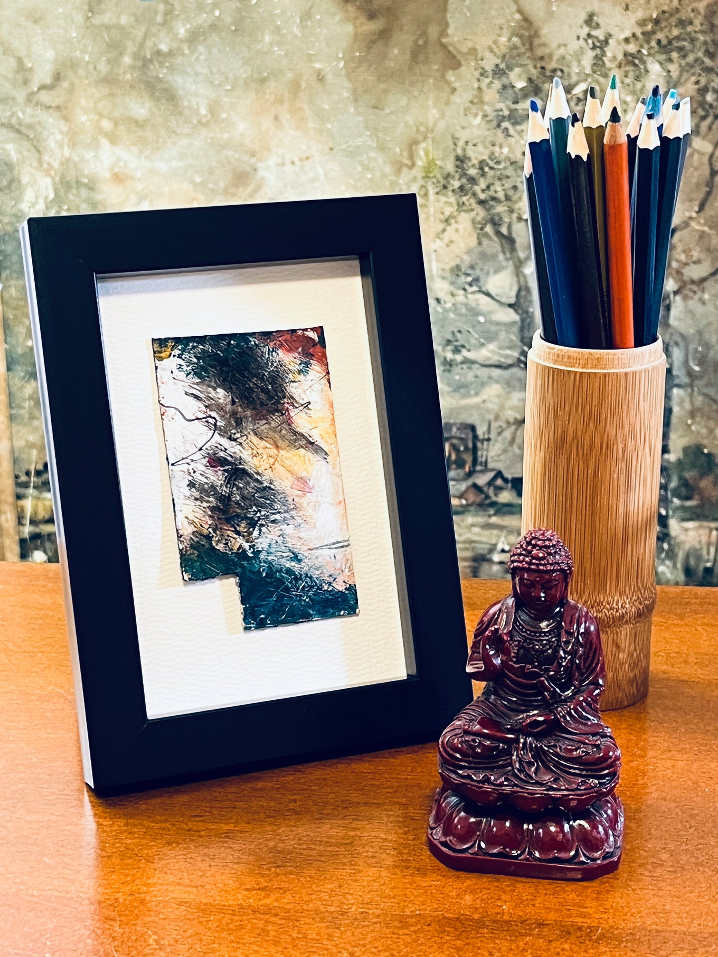 Original rectangular miniature abstract painting with a cut-out lower left corner revealing the mounting stock beneath. Painted in yellow, grey, peach, red, and teal colors of acrylic, ink, oil sticks and mascara on recycled packaging, text from the packaging is faintly visible. Mounted on 4x6-inch watercolor paper in portrait orientation, displayed in a 4x6-inch black frame next to a bamboo pencil holder and a small Buddha statue.