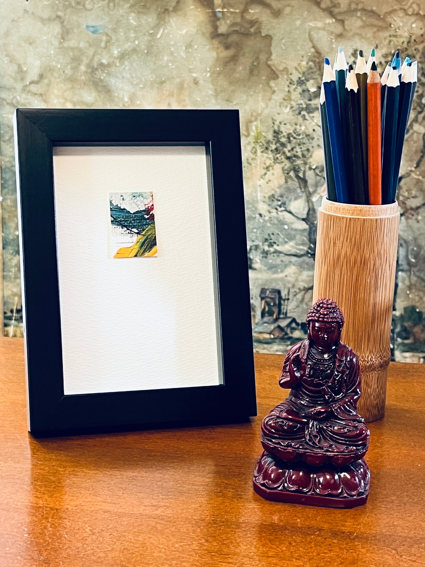 Original miniature abstract painting featuring yellow ochre, red and teal blue colors. Acrylic paint and ink on textured cold press watercolor paper, mounted on 4x6-inch watercolor paper in portrait orientation and displayed in a black frame next to a bamboo pencil holder and a small Buddha statue.