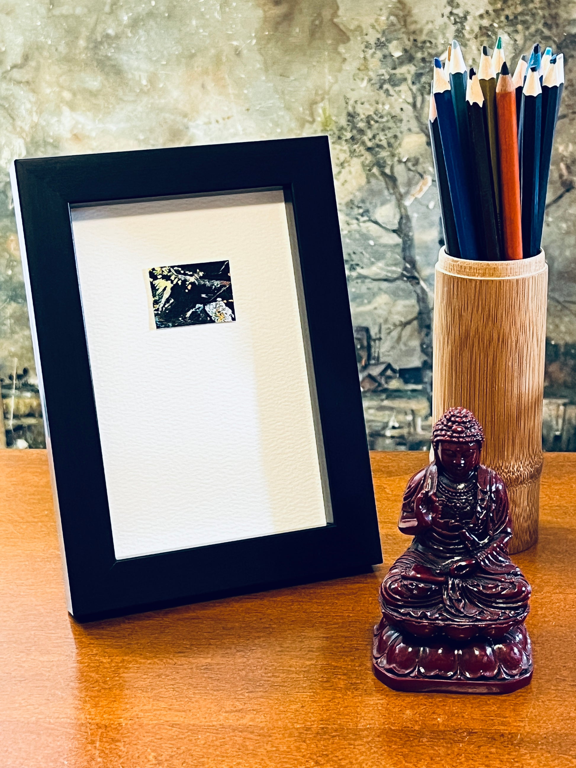 Original miniature abstract painting featuring yellow, white, orange and green colors. Acrylic paint on textured cold press watercolor paper, mounted on 4x6-inch watercolor paper in portrait orientation and displayed in a black frame next to a bamboo pencil holder and a small Buddha statue.