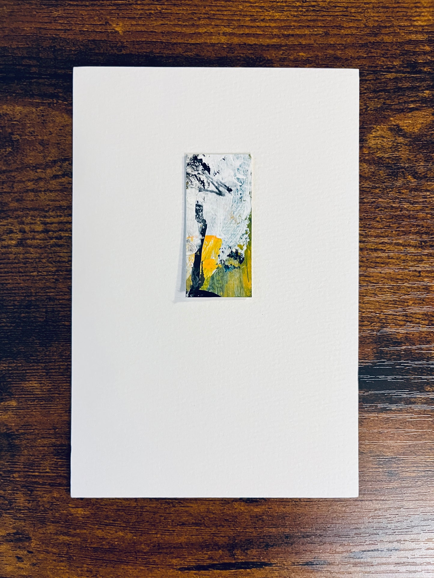 Original miniature abstract painting featuring yellow, green, white and black colors. Acrylic paint and ink on textured cold press watercolor paper, mounted on 4x6-inch watercolor paper in portrait orientation and photographed on a wooden table.