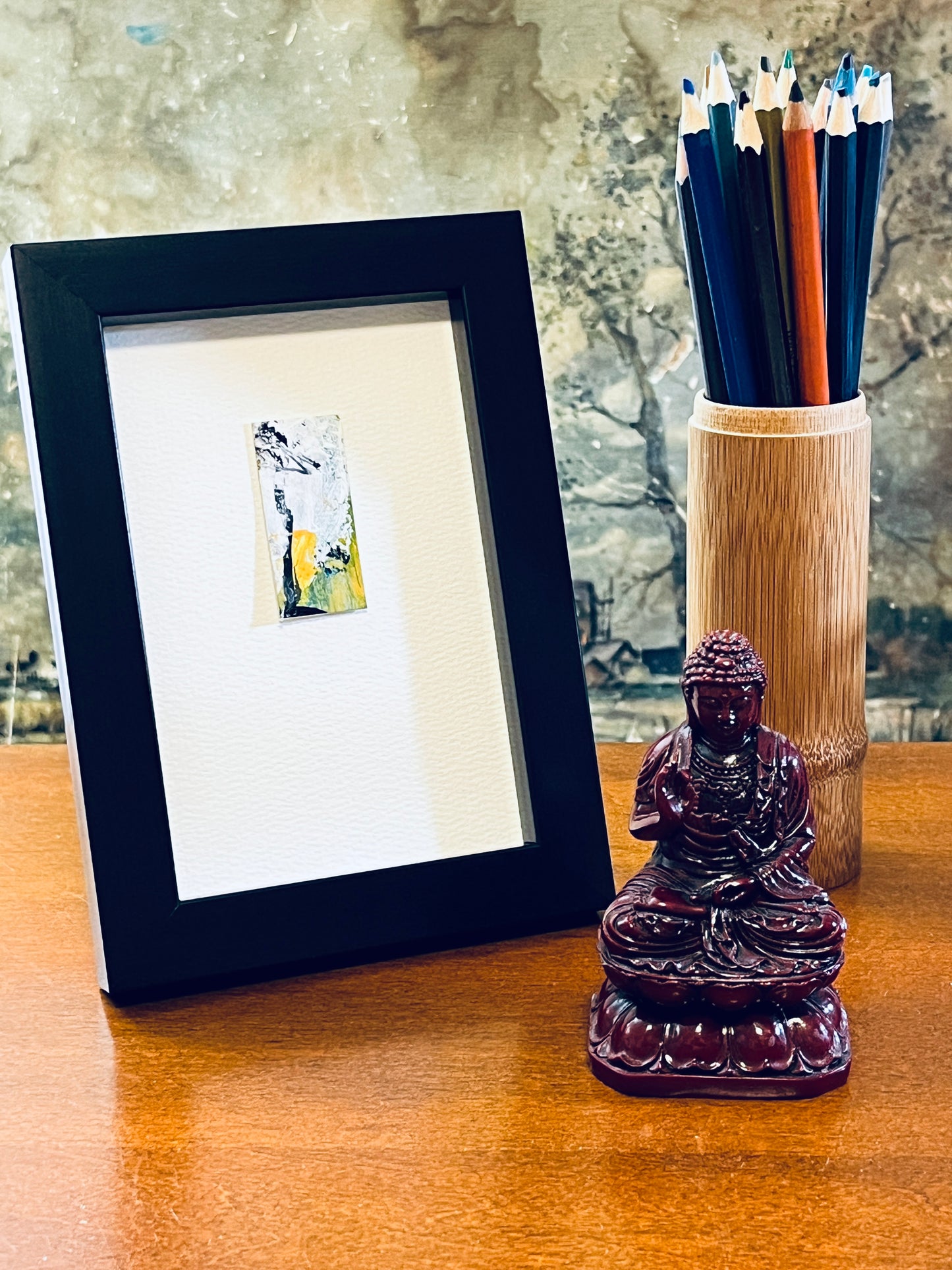 Original miniature abstract painting featuring yellow, green, white and black colors. Acrylic paint and ink on textured cold press watercolor paper, mounted on 4x6-inch watercolor paper in portrait orientation displayed in a black frame next to a bamboo pencil holder and a small Buddha statue..