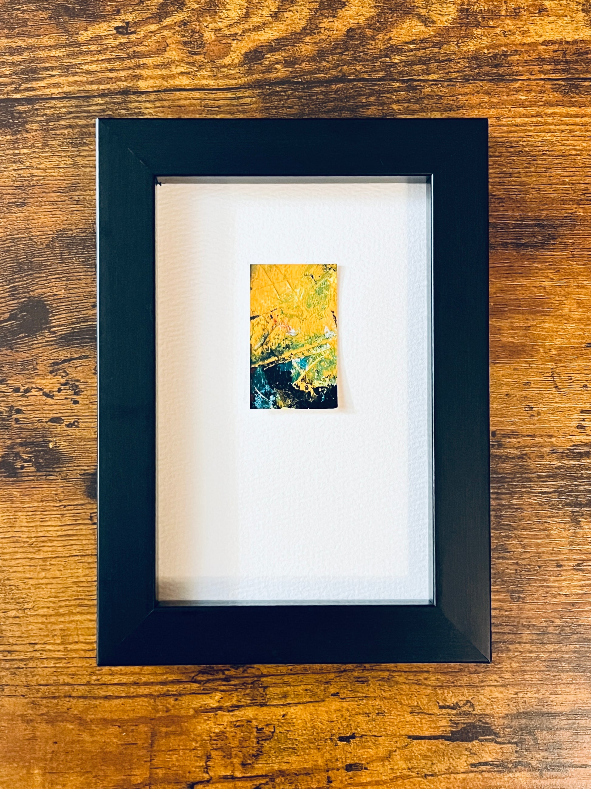 Original miniature abstract painting featuring yellow ochre, gree, black and red colors. Acrylic paint on textured cold press watercolor paper, mounted on 4x6-inch watercolor paper in portrait orientation and photographed in a black 4x6-inch frame on a wooden table.