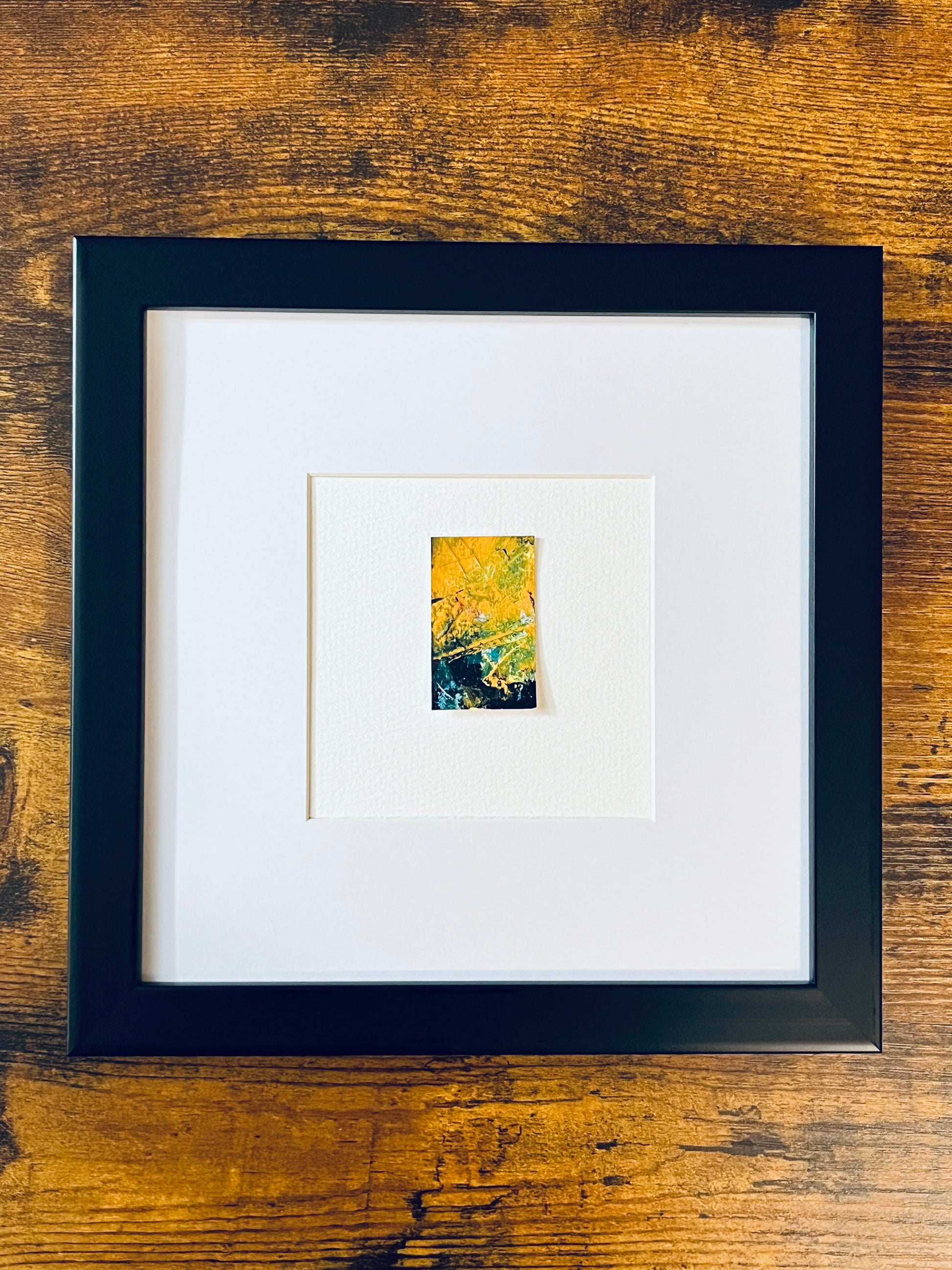 Original miniature abstract painting featuring yellow ochre, gree, black and red colors. Acrylic paint on textured cold press watercolor paper, mounted on 4x6-inch watercolor paper in portrait orientation and photographed in a black 8x8-inch frame on a wooden table.