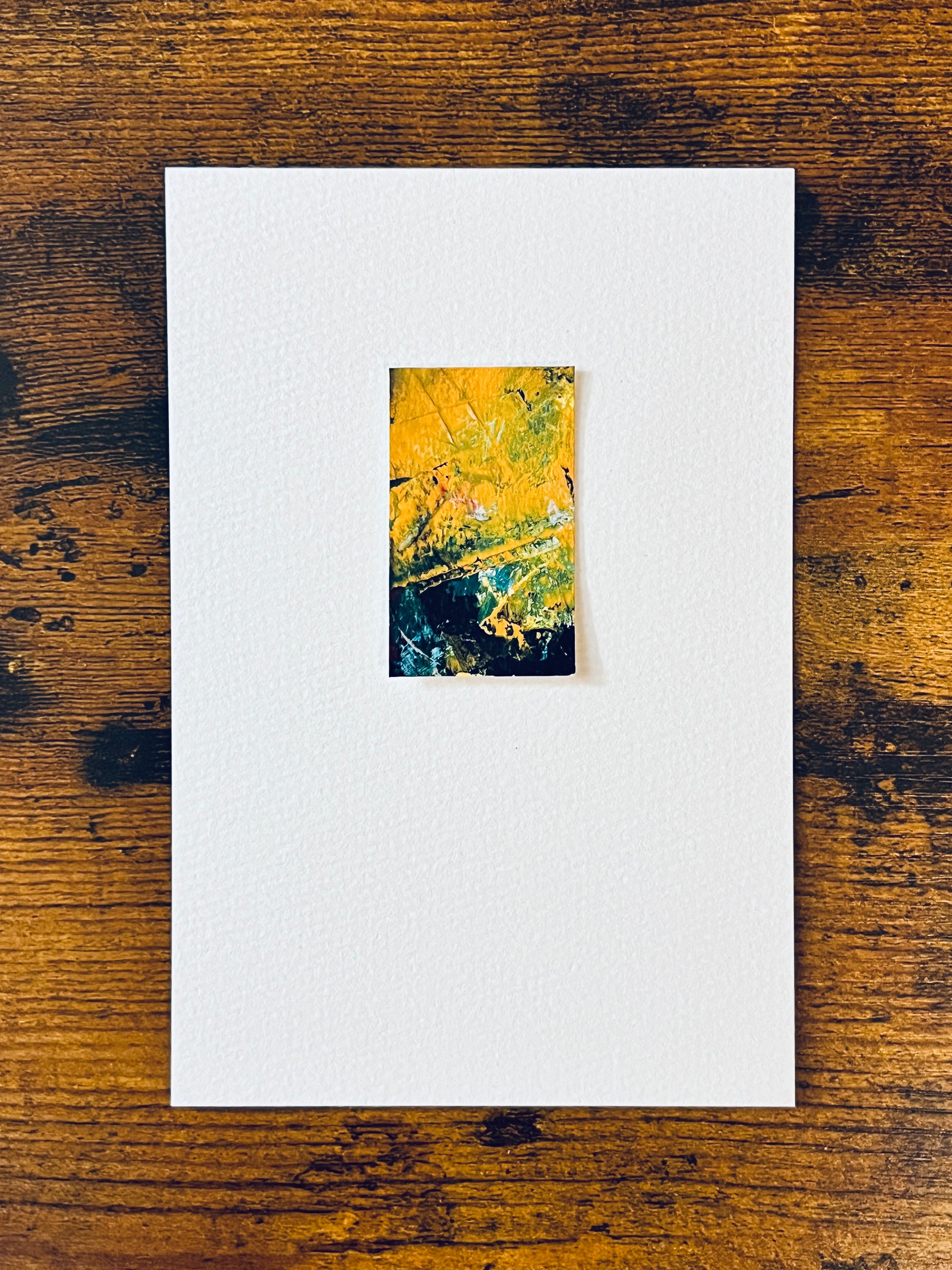 Original miniature abstract painting featuring yellow ochre, gree, black and red colors. Acrylic paint on textured cold press watercolor paper, mounted on 4x6-inch watercolor paper in portrait orientation and photographed on a wooden table.