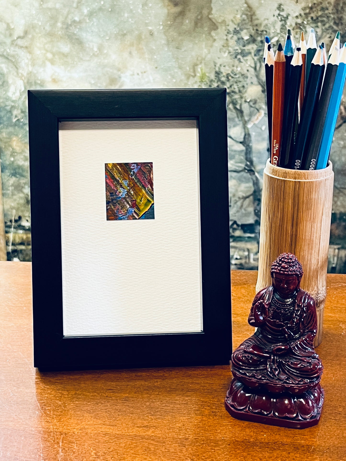 Original miniature abstract painting featuring yellow ochre, red, pink, and blue colors. Acrylic paint on textured cold press watercolor paper, mounted on 4x6-inch watercolor paper in portrait orientation and displayed in a black frame next to a bamboo pencil holder and a small Buddha statue.