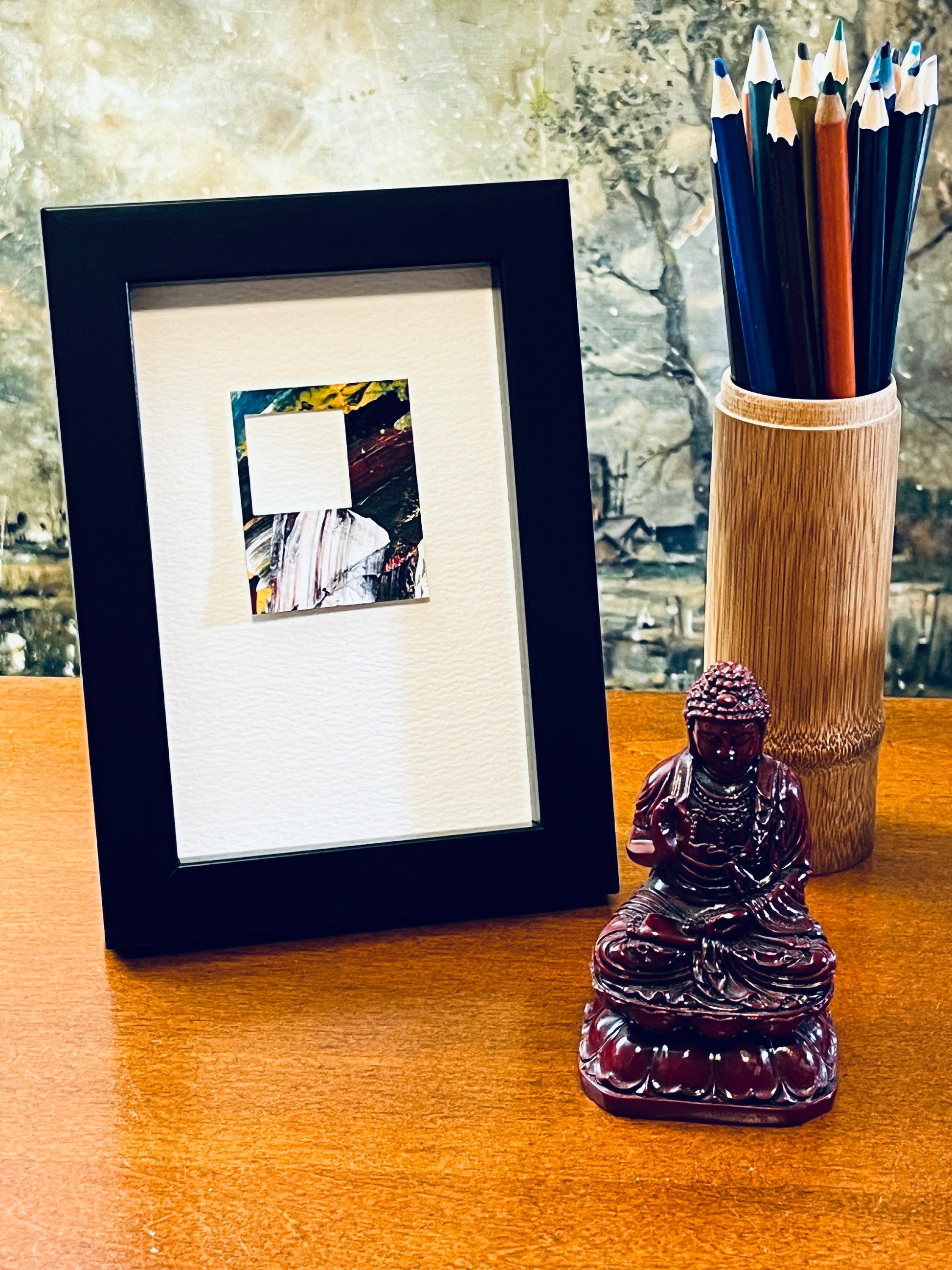 Original miniature abstract painting with a window cutout in the upper left revealing the mounting stock. Painted with acrylic paint in yellow, red and teal blue colors on textured cold press watercolor paper, mounted on 4x6-inch watercolor paper in portrait orientation and displayed in a black frame next to a bamboo pencil holder and a small Buddha statue.