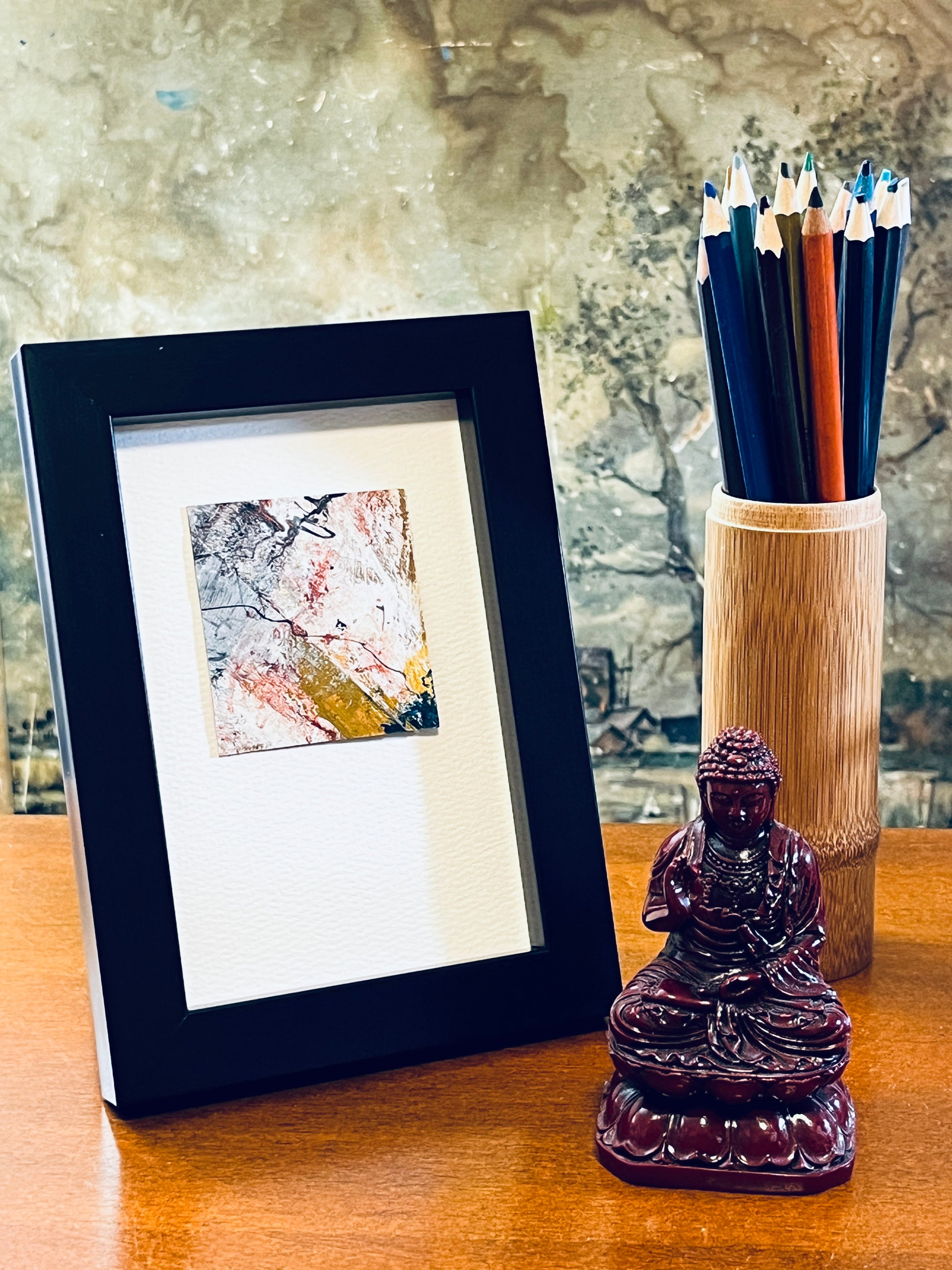 Original miniature abstract painting in yellow ochre, grey, peach, red, and teal colors of acrylic, ink, oil sticks and mascara on recycled packaging with a visible diagonal seam incorporated into the painting. Mounted on 4x6-inch watercolor paper in portrait orientation,displayed in a black frame next to a bamboo pencil holder and a small Buddha statue.