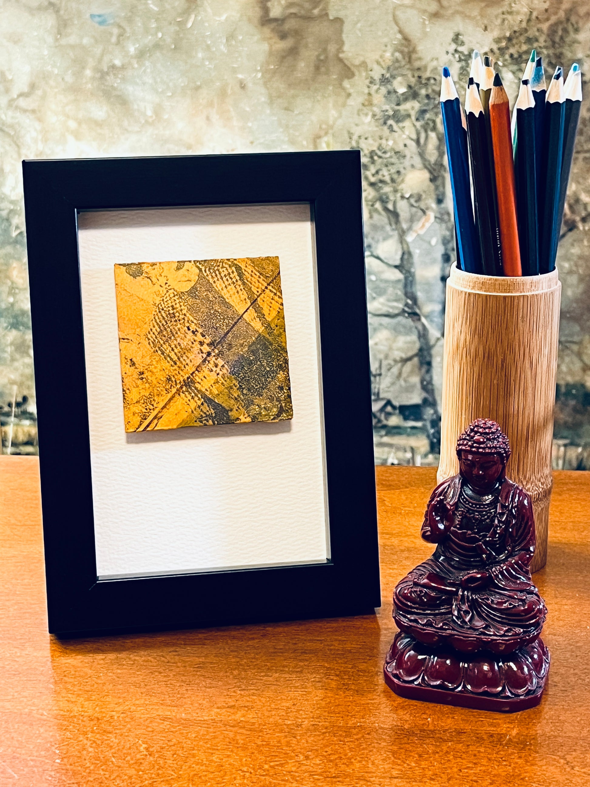 Original miniature abstract monoprint painted envelope in yellow ochre and grey colors. Acrylic paint on fine-grain bond paper, mounted on 4x6-inch watercolor paper in portrait orientation and displayed in a black frame next to a bamboo pencil holder and a small Buddha statue.
