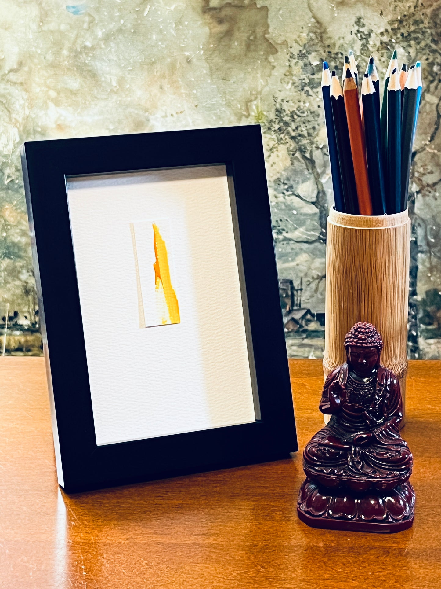 Original miniature abstract painting featuring yellow ochre acrylic paint on textured cold press watercolor paper, mounted on 4x6-inch watercolor paper in portrait orientation and displayed in a black frame next to a bamboo pencil holder and a small Buddha statue.