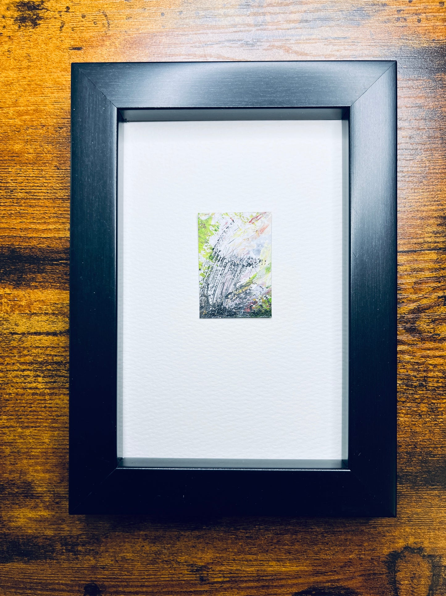 Original artwork by Anet Skillin. This miniature abstract painting is painted on recycled packaging in yellow-green, grey and white and red colors; mounted on 4x6-inch paper and photographed in a black 4x6-inch frame against a wooden table.