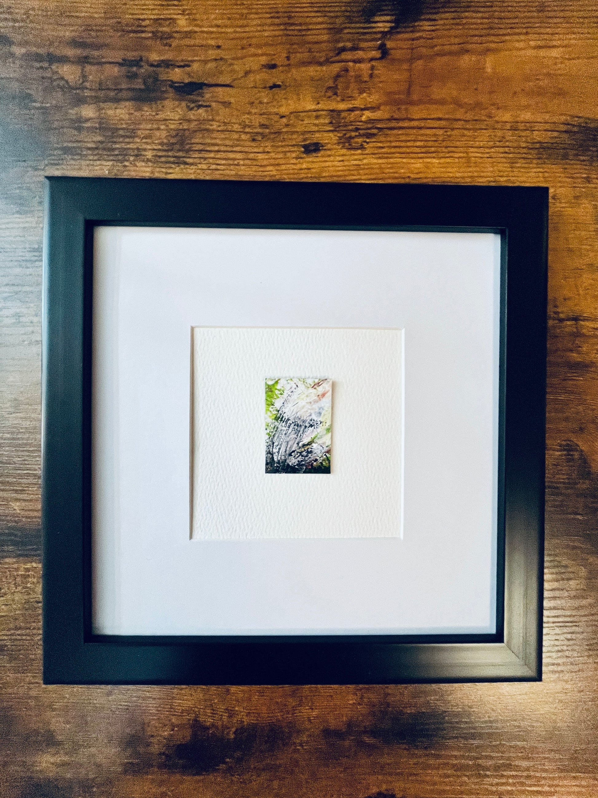 Original artwork by Anet Skillin. This miniature abstract painting is painted on recycled packaging in yellow-green, grey and white and red colors; mounted on 4x6-inch paper and photographed in a black 8x8-inch frame against a wooden table.