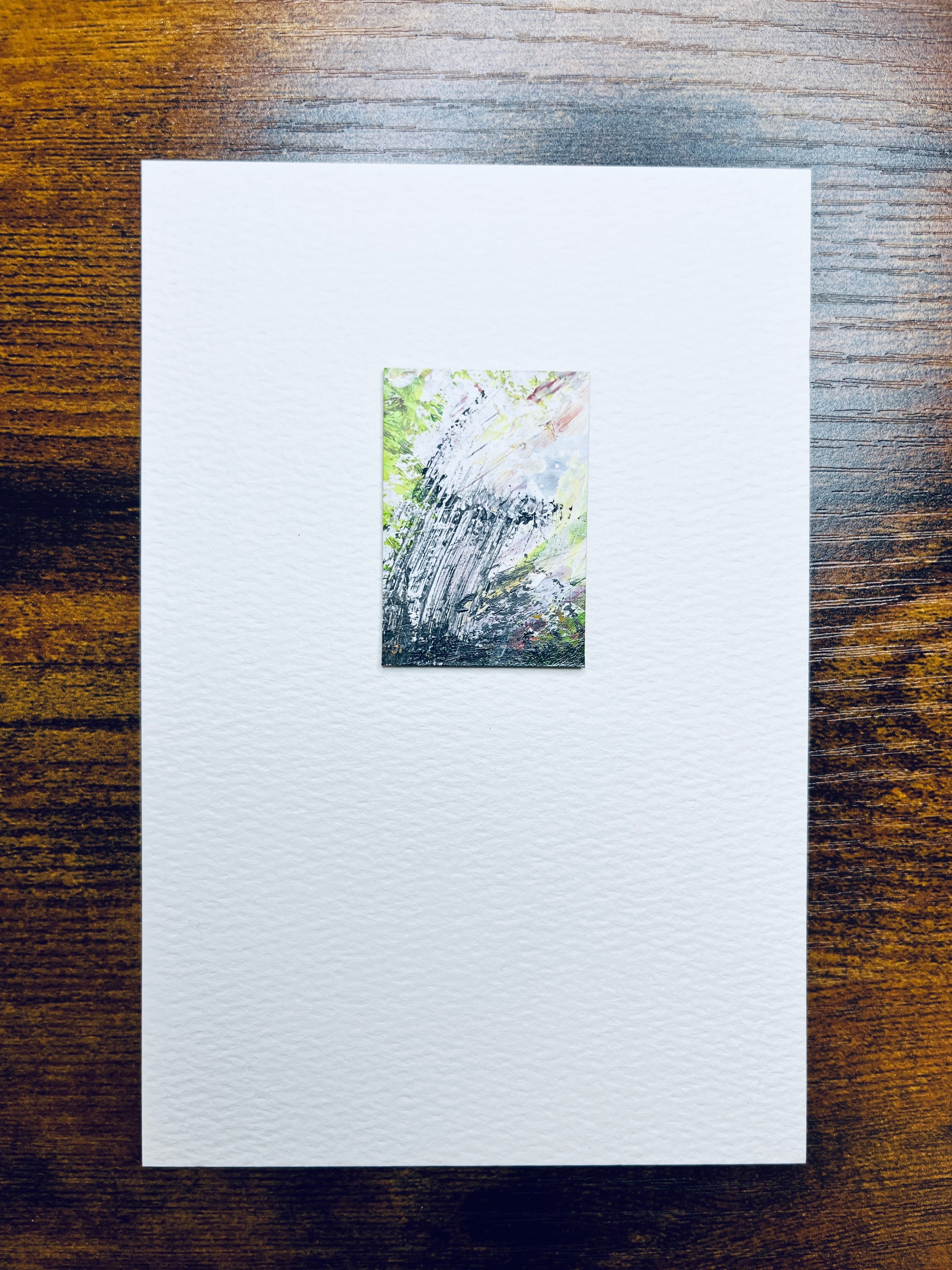 Original artwork by Anet Skillin. This miniature abstract painting is painted on recycled packaging in yellow-green, grey and white colors; mounted on 4x6-inch paper and photographed against a wooden table.