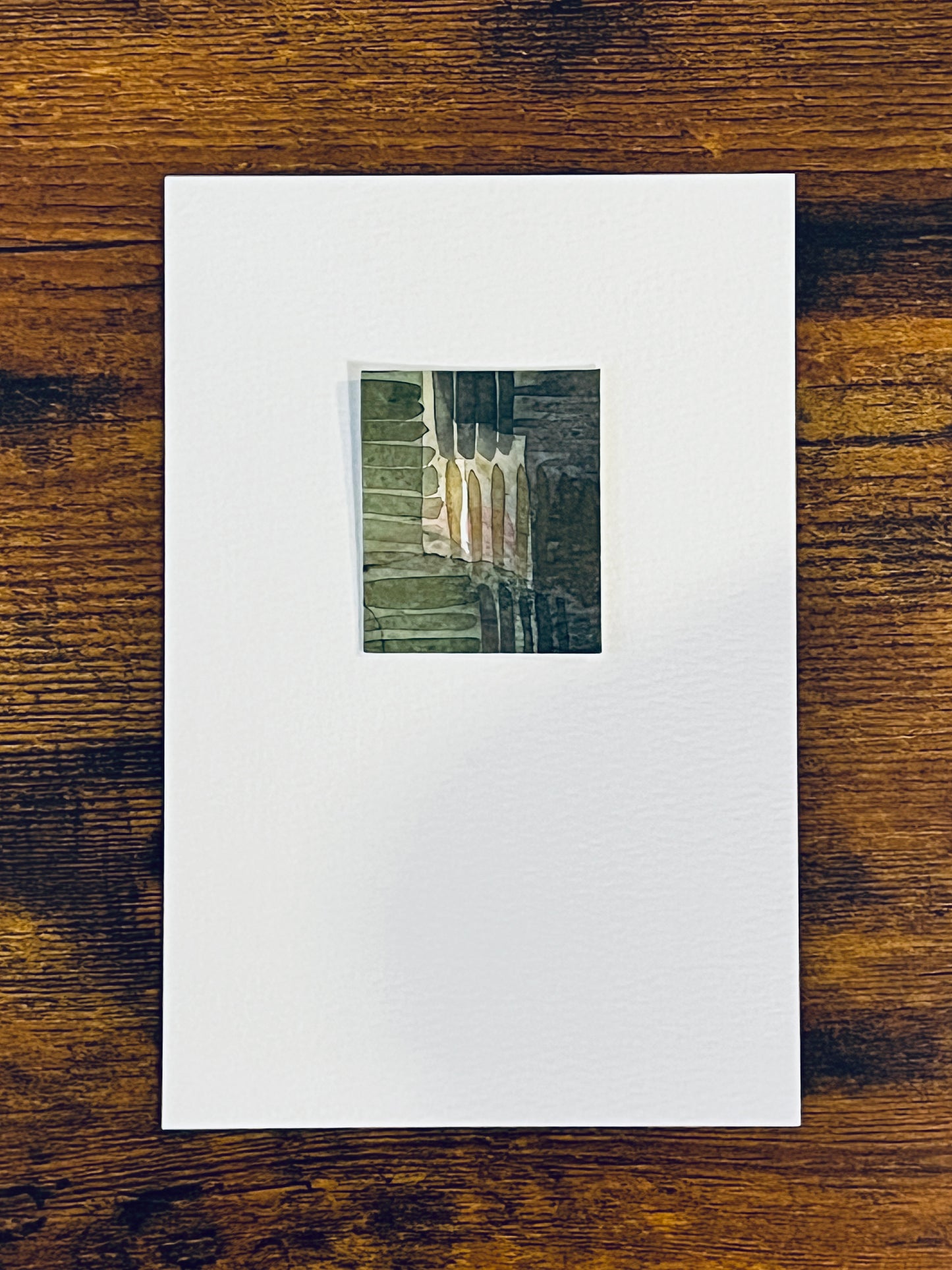 Original miniature abstract painting of plaid fabric featuring green and grey shades with yellow ochre and some pink. Watercolor paint on textured cold press watercolor paper, mounted on 4x6-inch watercolor paper in portrait orientation and photographed on a wooden table.