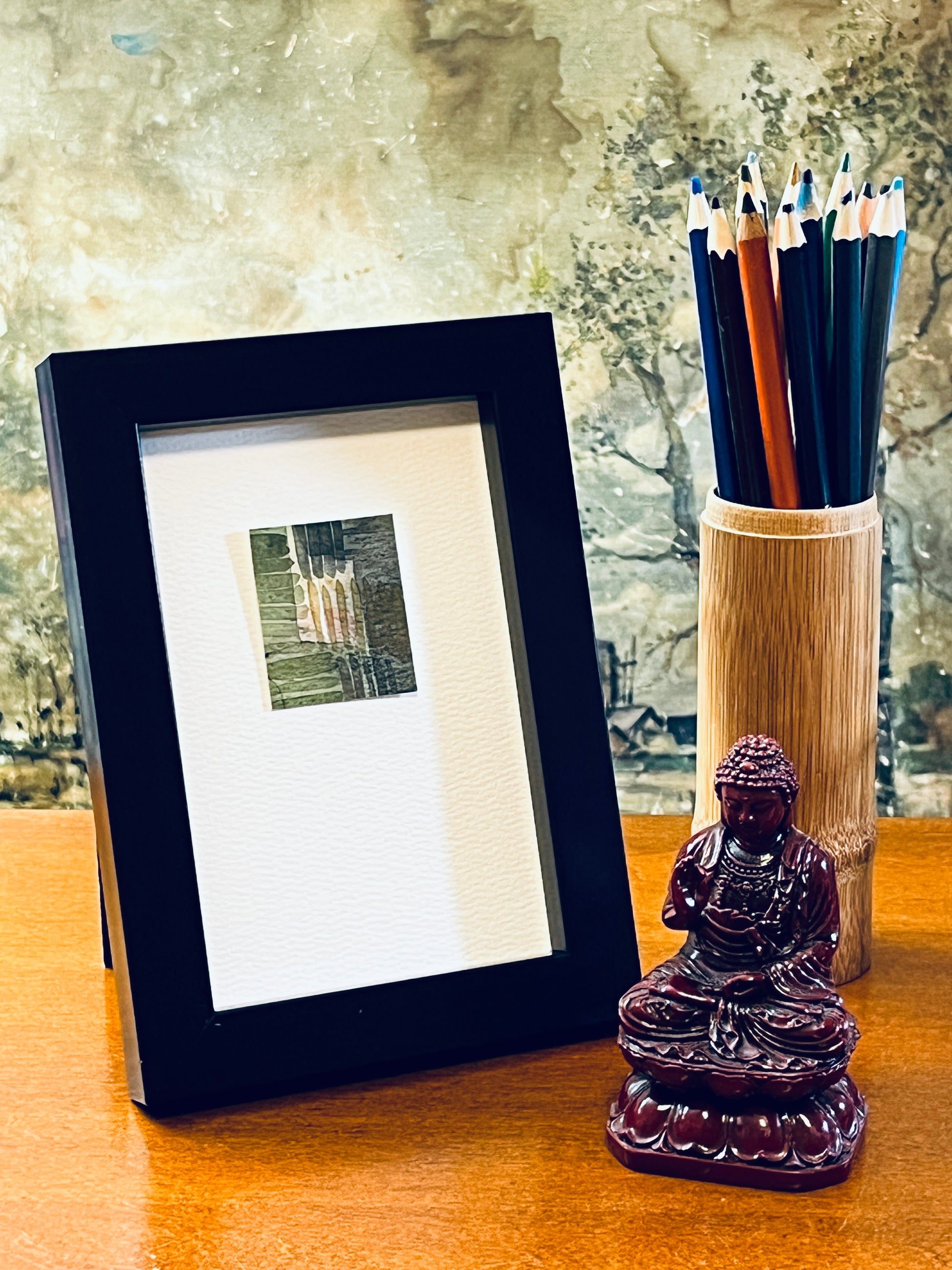 Original miniature abstract painting of plaid fabric featuring green and grey shades with yellow ochre and some pink. Watercolor paint on textured cold press watercolor paper, mounted on 4x6-inch watercolor paper in portrait orientation and displayed in a black frame next to a bamboo pencil holder and a small Buddha statue.