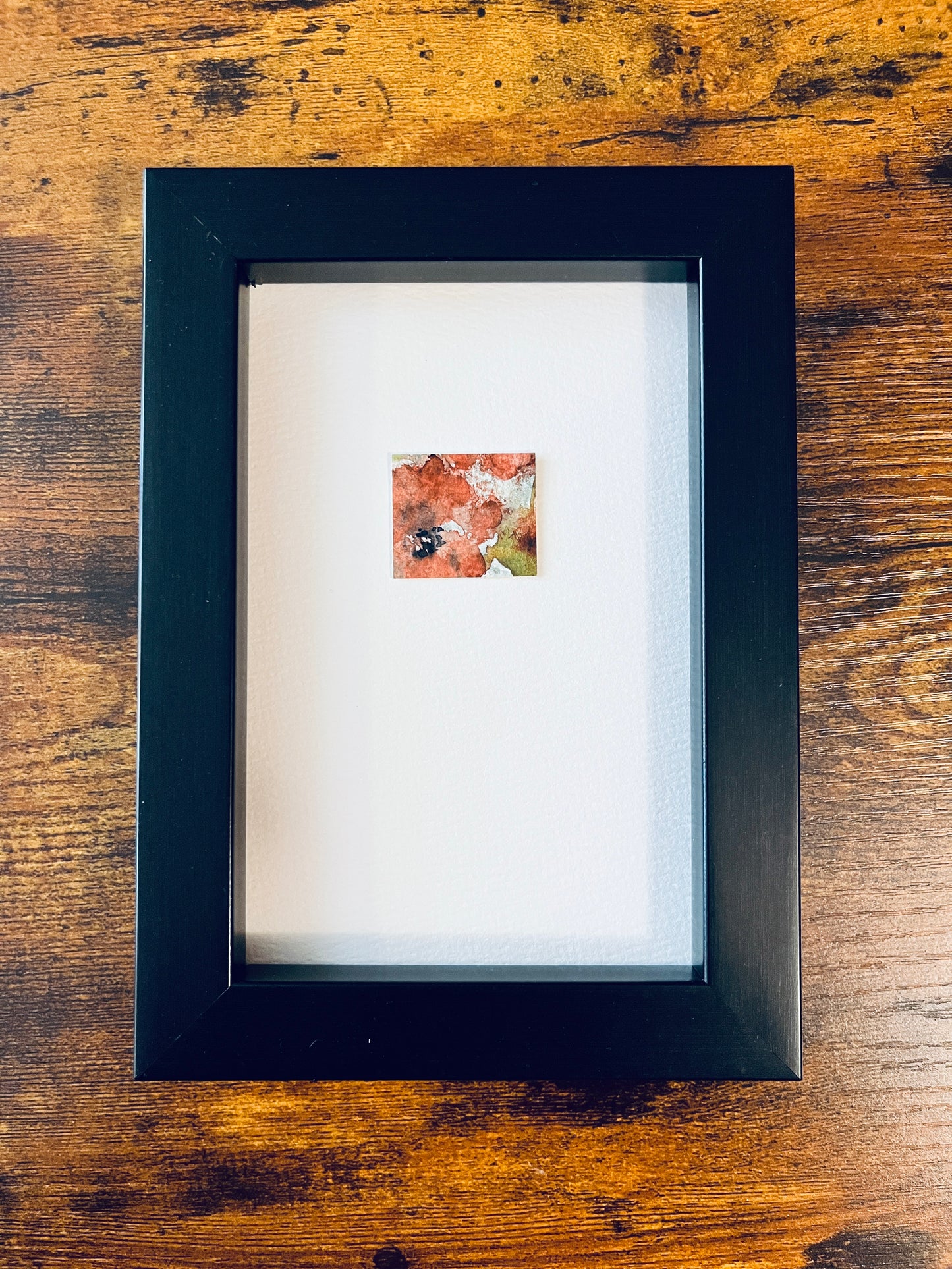 Original artwork by Anet Skillin. This miniature abstract floral painting features a poppy. Painted on textured cold-press watercolor paper, in orange-red, yellow-green, pale blue and black colors; mounted on 4x6-inch paper and photographed in a black 4x6-inch frame against a wooden table.