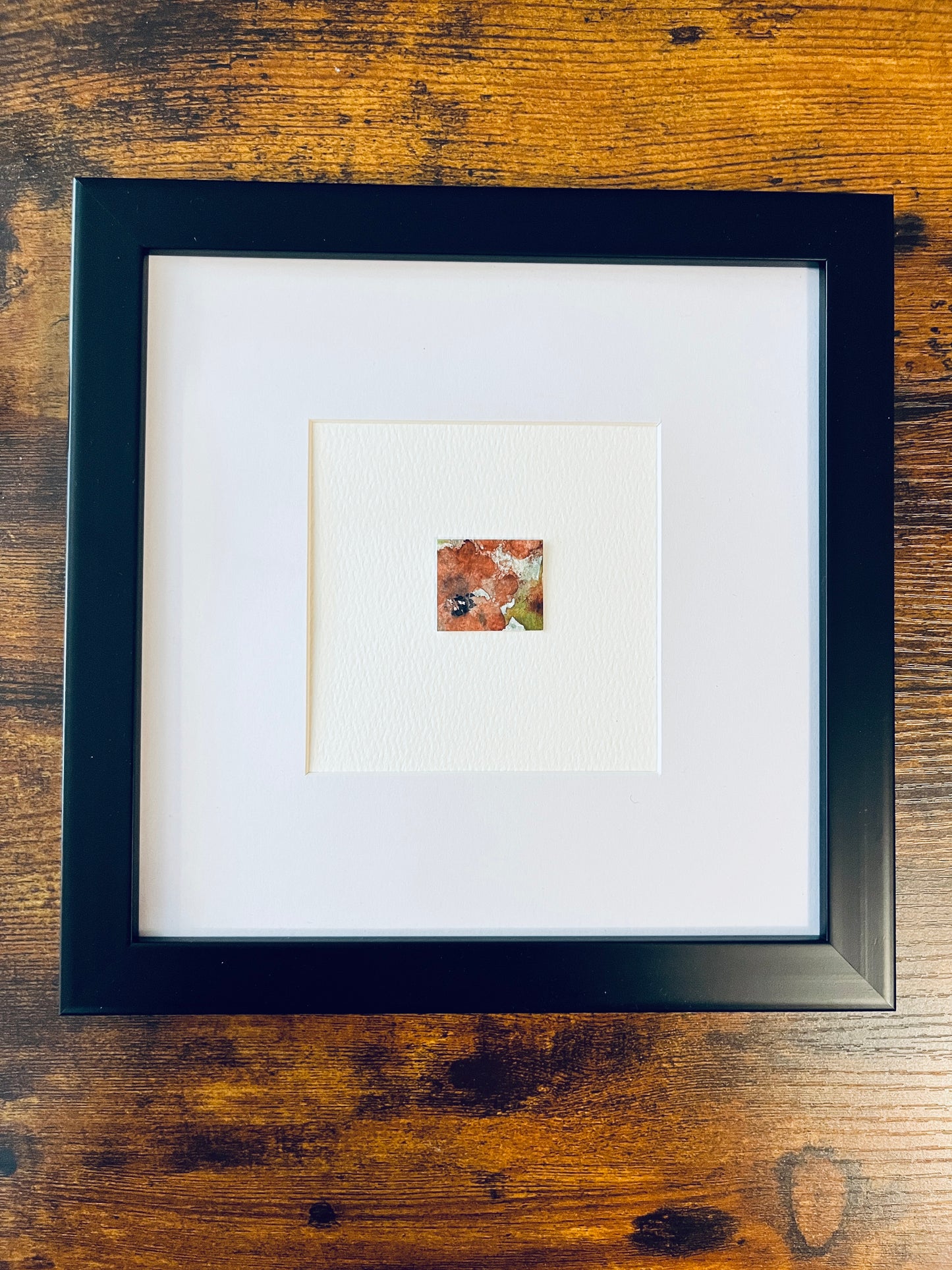 Original artwork by Anet Skillin. This miniature abstract floral painting features a poppy. Painted on textured cold-press watercolor paper, in orange-red, yellow-green, pale blue and black colors; mounted on 4x6-inch paper and photographed in a black 8x8-inch frame against a wooden table.