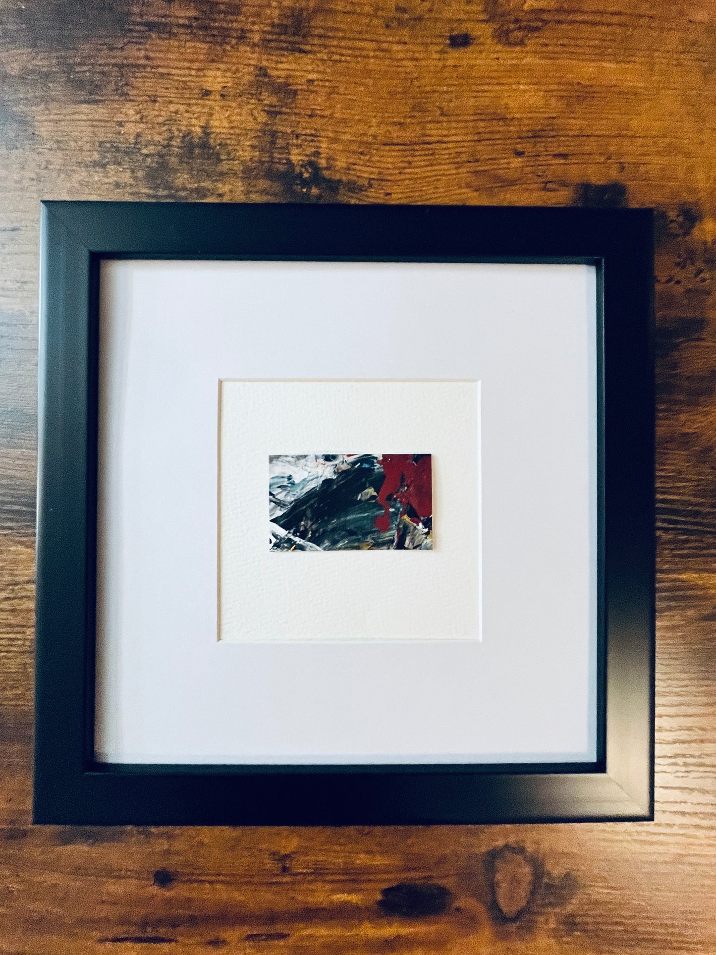 Original artwork by Anet Skillin. This miniature abstract painting is painted on Kraft paper, in black, white, red, blue and yellow ochre colors; mounted on 4x6-inch paper and photographed in a 8x8-inch black frame against a wooden table.