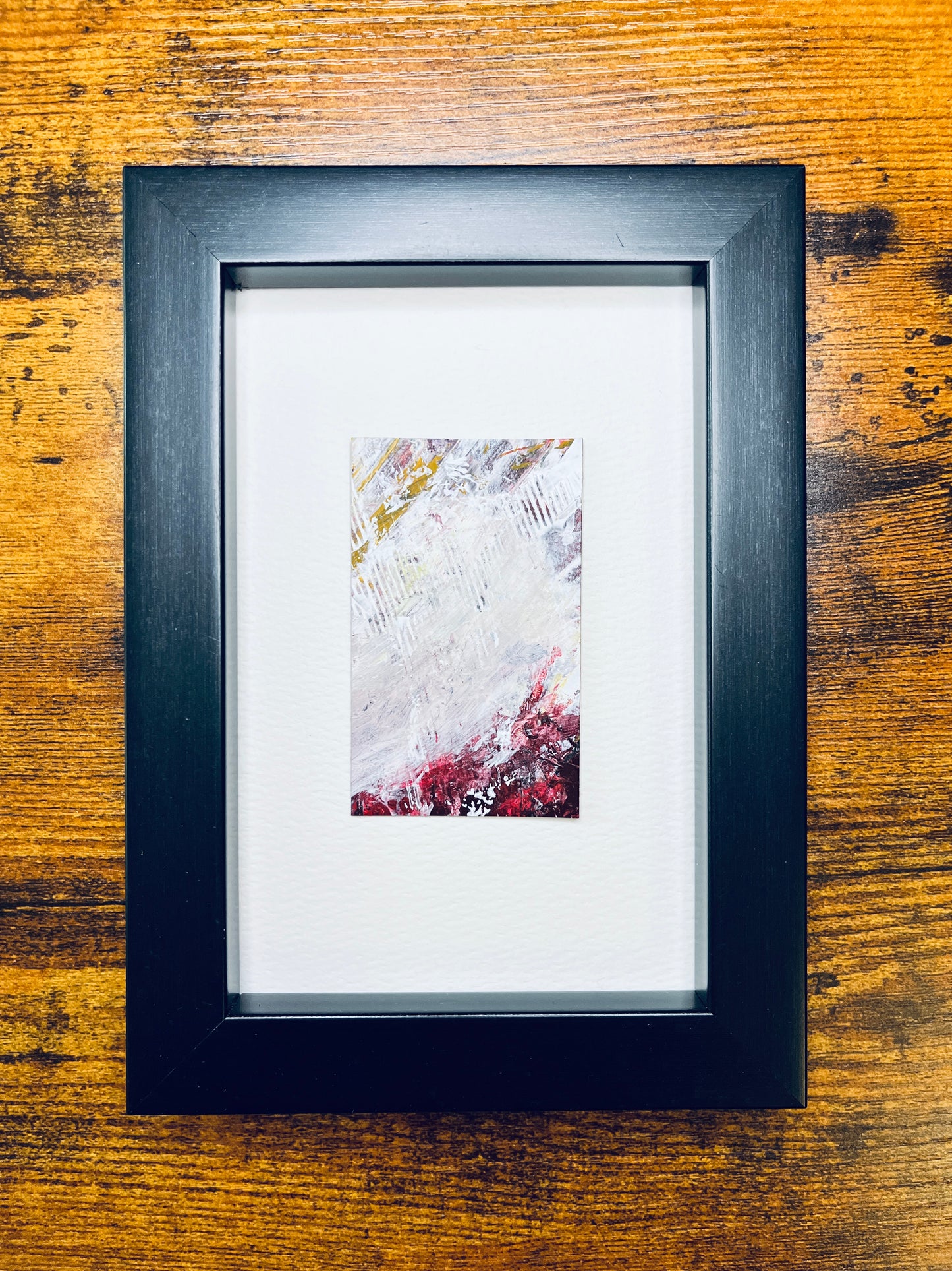 Original artwork by Anet Skillin. This miniature abstract is painted on recycled packaging in white, red, yellow-ochre and grey colors; mounted on 4x6-inch paper and photographed in a black 4x6-inch frame against a wooden table.