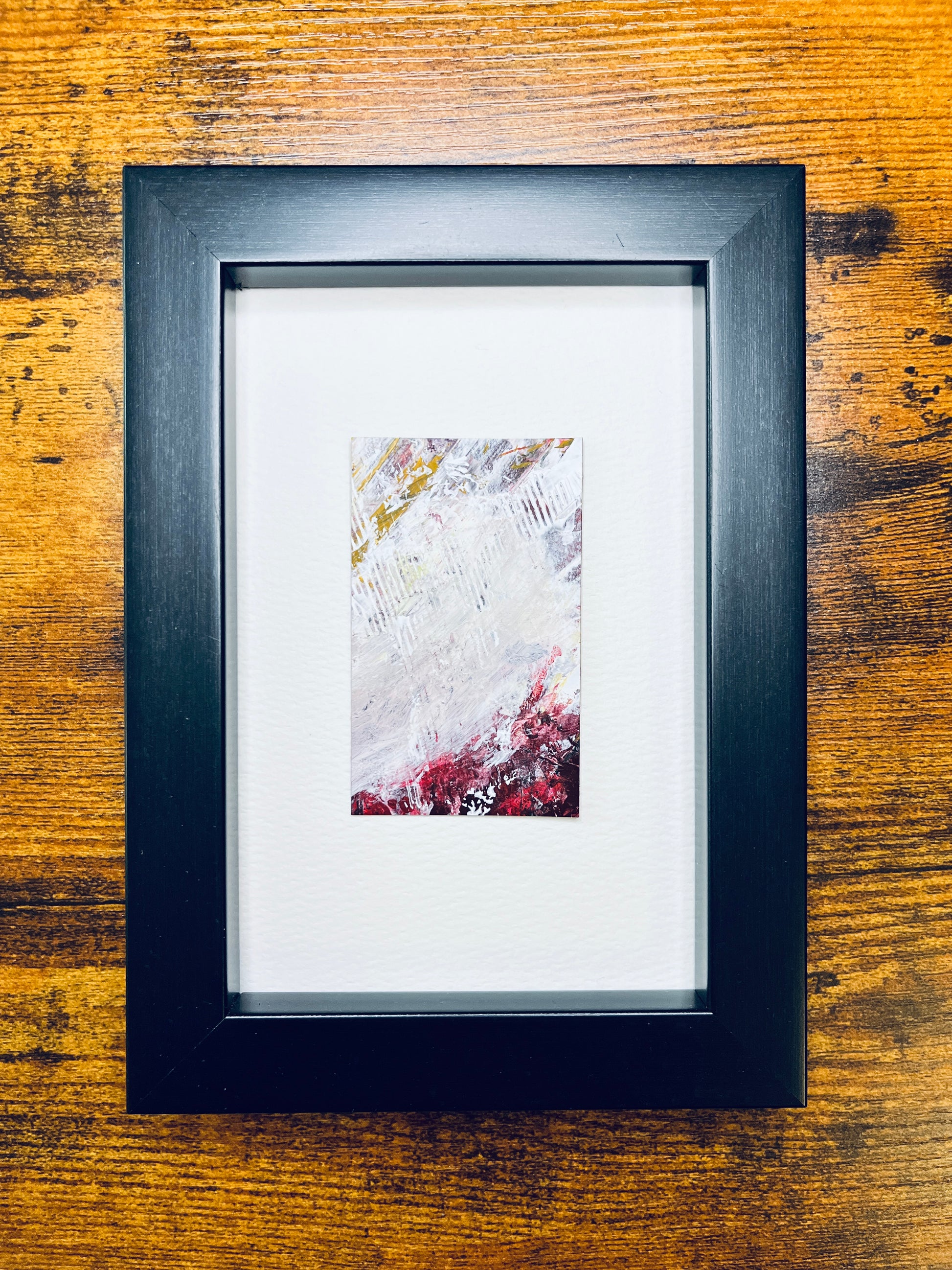 Original artwork by Anet Skillin. This miniature abstract is painted on recycled packaging in white, red, yellow-ochre and grey colors; mounted on 4x6-inch paper and photographed in a black 4x6-inch frame against a wooden table.