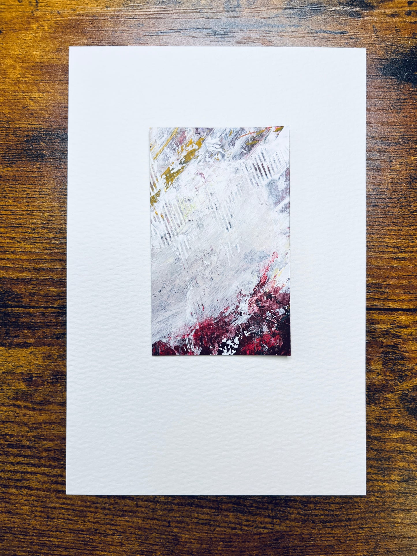 Original artwork by Anet Skillin. This miniature abstract is painted on recycled packaging in white, red, yellow-ochre and grey colors; mounted on 4x6-inch paper and photographed against a wooden table.