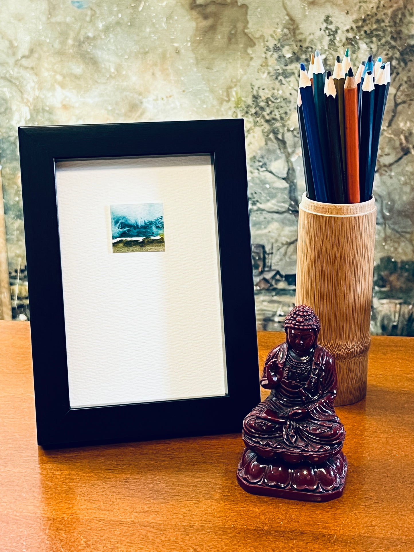 Original miniature abstract landscape painting featuring blue-green, brown and green colors. Watercolor paint textured cold press watercolor paper, mounted on 4x6-inch watercolor paper in portrait orientation and displayed in a black frame next to a bamboo pencil holder and a small Buddha statue.