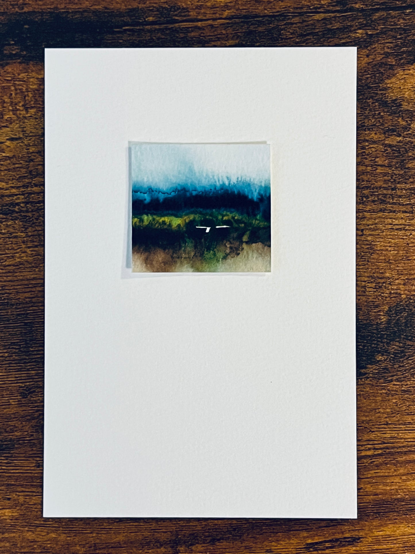 Original miniature abstract landscape painting featuring watery blues, brown, green and yellow ochre colors. Watercolor paint on textured cold press watercolor paper, mounted on 4x6-inch watercolor paper in portrait orientation and photographed on a wooden table.