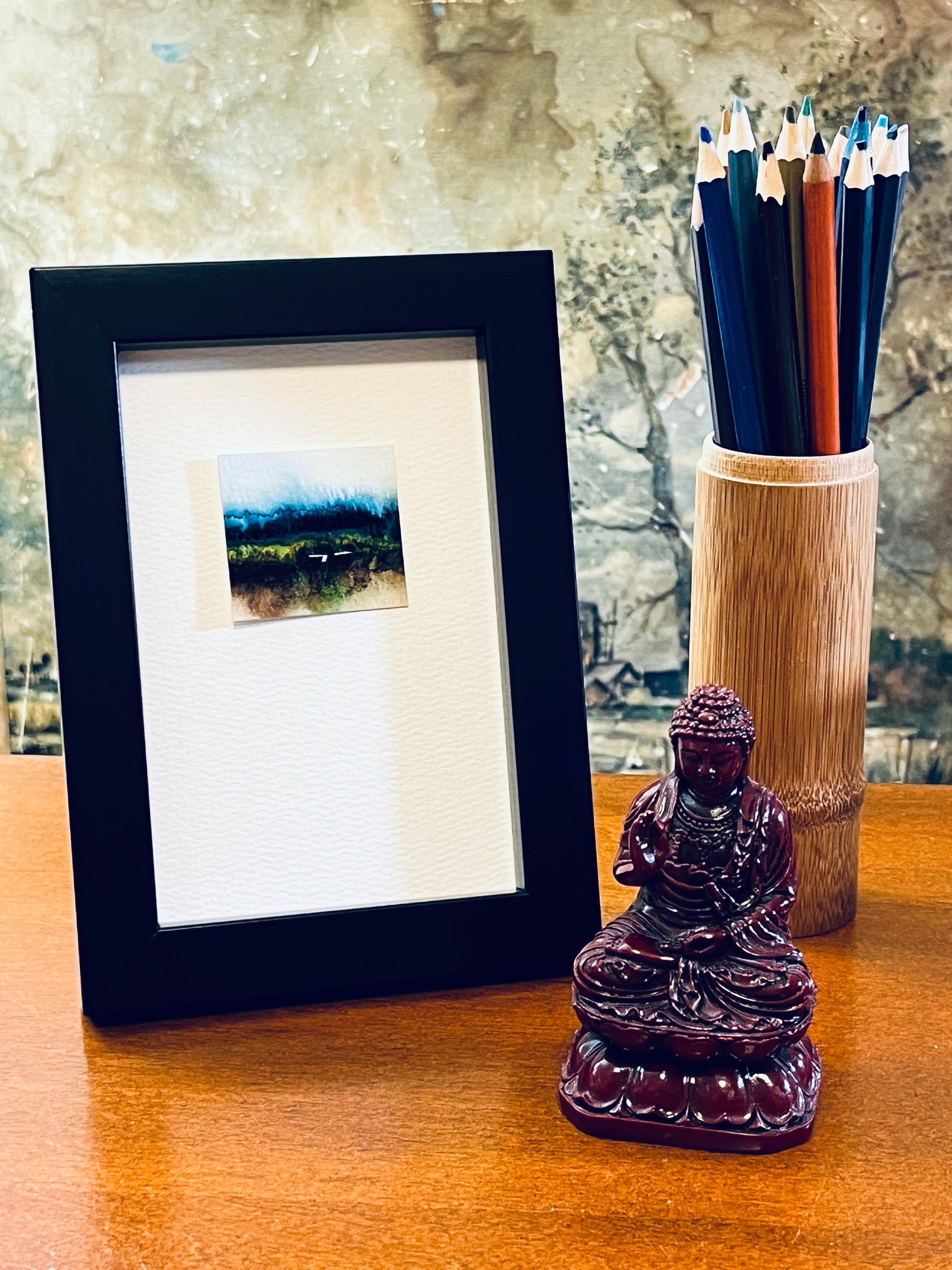 Original miniature abstract landscape painting featuring watery blues, brown, green and yellow ochre colors. Watercolor paint on textured cold press watercolor paper, mounted on 4x6-inch watercolor paper in portrait orientation displayed in a black frame next to a bamboo pencil holder and a small Buddha statue.