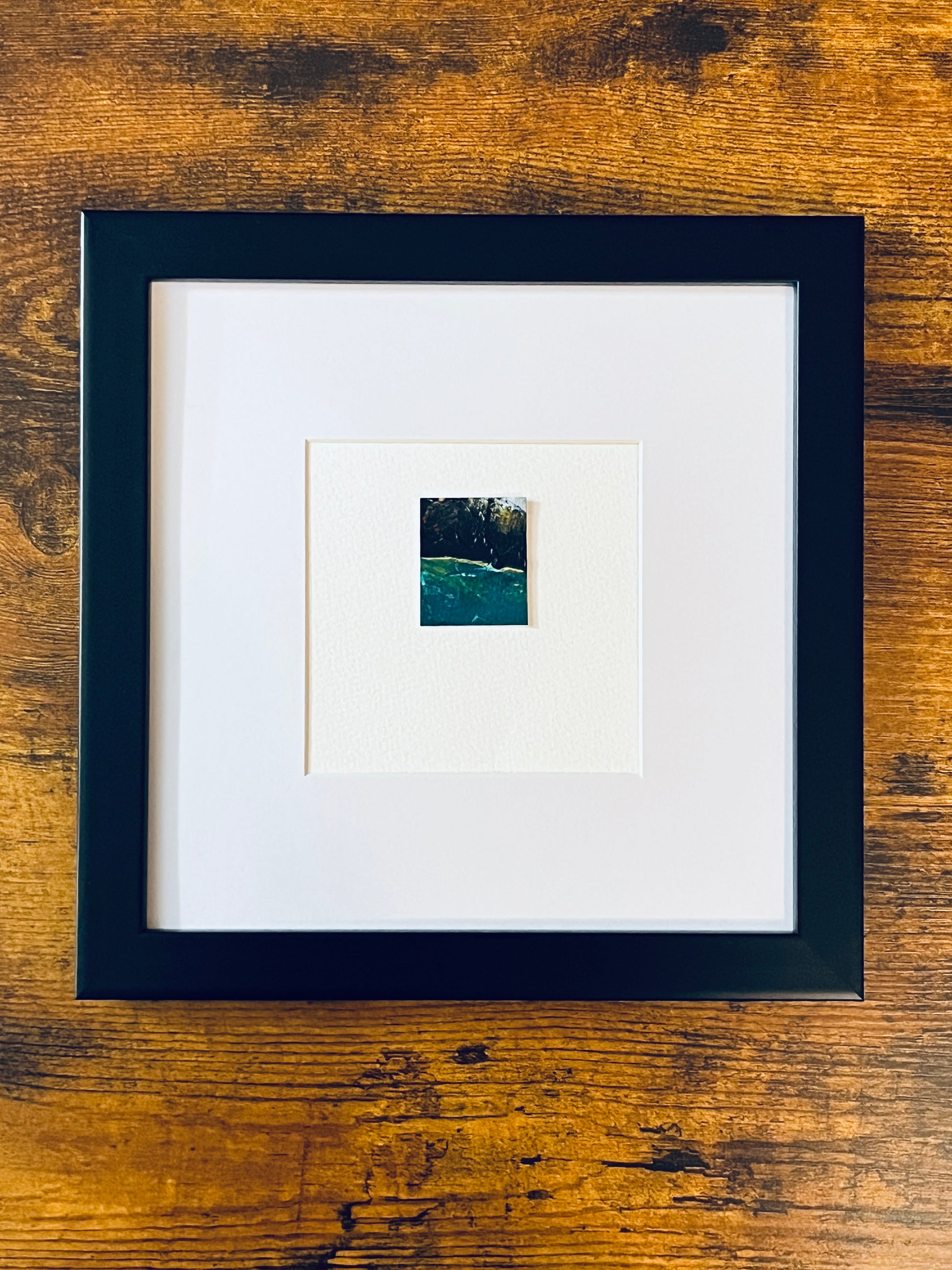 Original miniature abstract painting featuring green, yellow, red and black colors. Acrylic paint on textured cold press watercolor paper, mounted on 4x6-inch watercolor paper in portrait orientation and photographed in a black 8x8-inch frame on a wooden table.