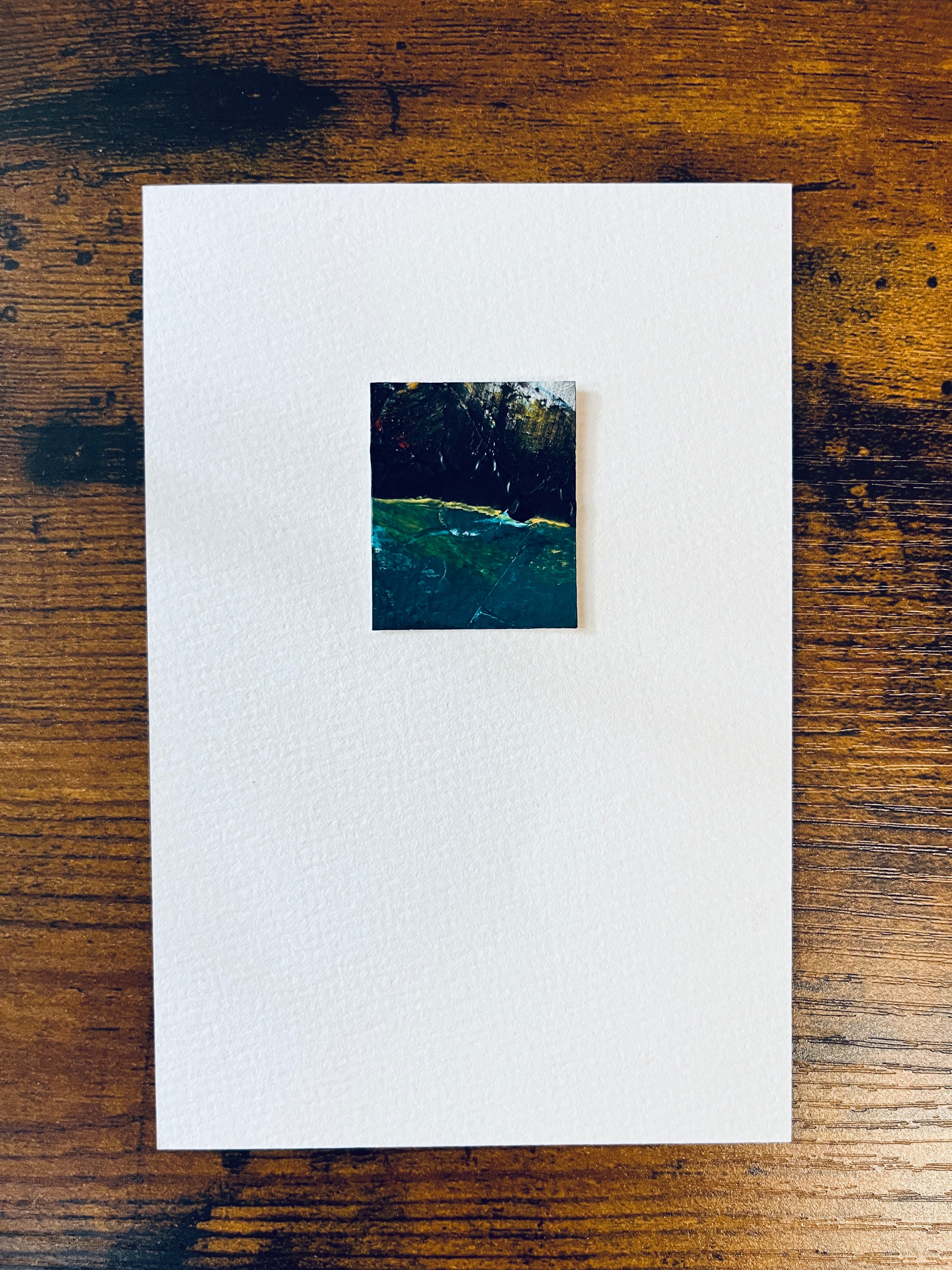 Original miniature abstract painting featuring green, yellow, red and black colors. Acrylic paint on textured cold press watercolor paper, mounted on 4x6-inch watercolor paper in portrait orientation and photographed on a wooden table.