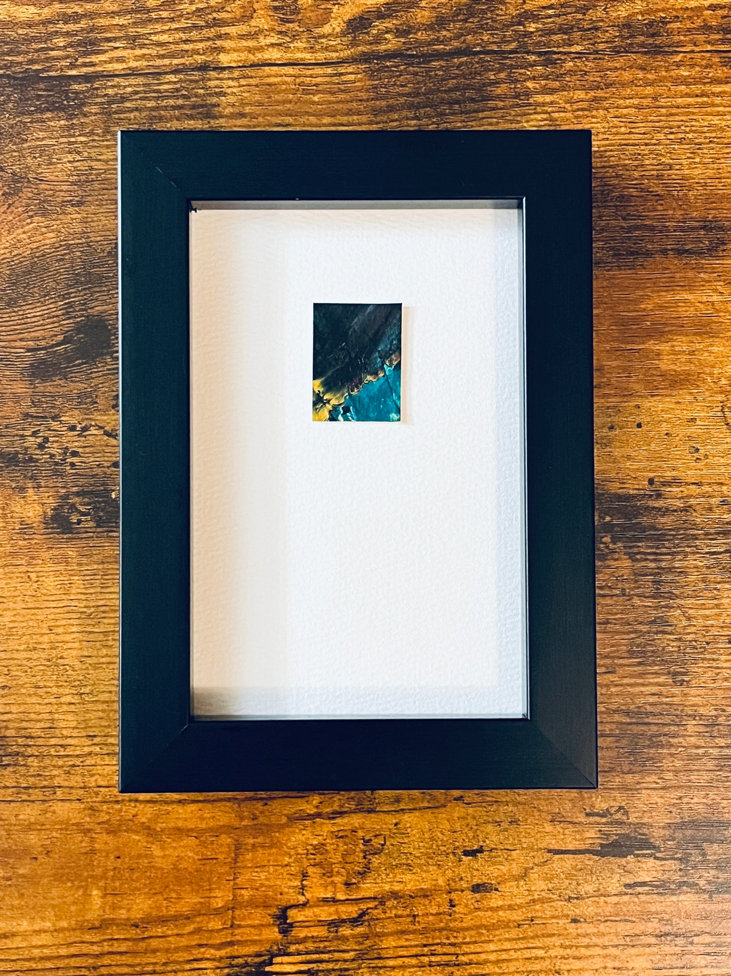 Original miniature abstract painting featuring green, black and yellow ochre colors. Acrylic paint on textured cold press watercolor paper, mounted on 4x6-inch watercolor paper in portrait orientation and photographed in a black 4x6-inch frame on a wooden table.