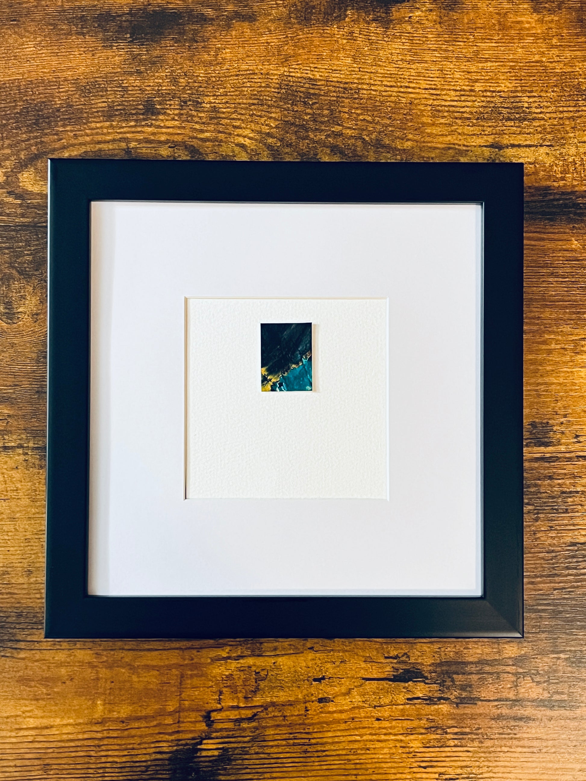 Original miniature abstract painting featuring green, black and yellow ochre colors. Acrylic paint on textured cold press watercolor paper, mounted on 4x6-inch watercolor paper in portrait orientation and photographed in a black 8x8-inch frame on a wooden table.