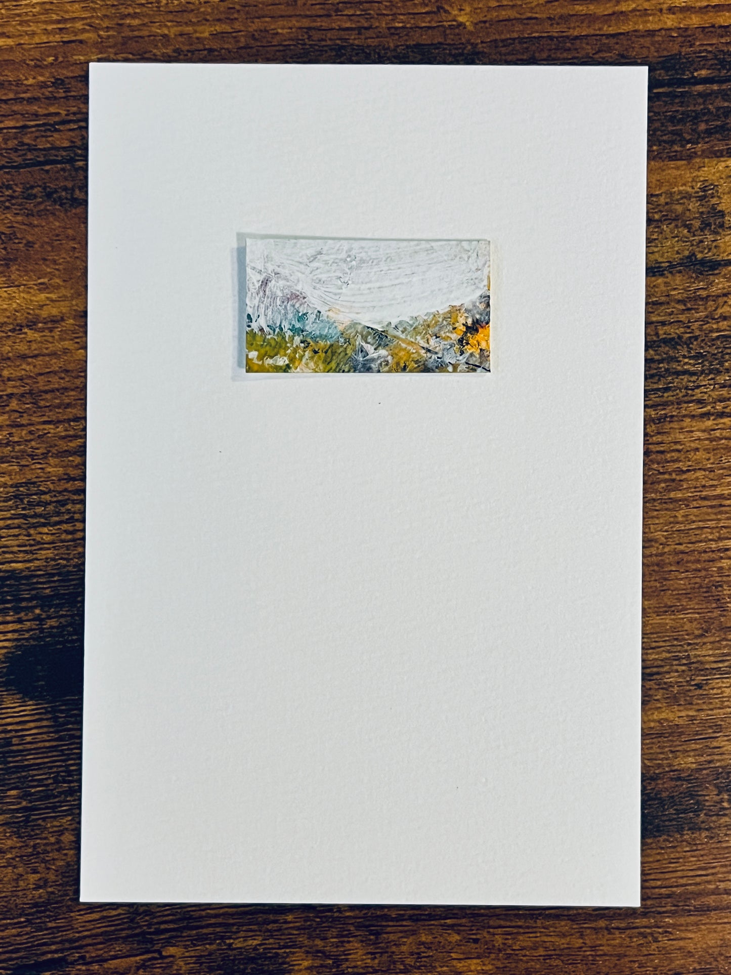 Original miniature abstract landscape painting featuring a mountain scene in spring foliage with a snowy backdrop. White, turquoise, yellow ochre, mint green orange and a touch of red is used. Acrylic paint on textured cold press watercolor paper, mounted on 4x6-inch watercolor paper in portrait orientation and photographed on a wooden table.