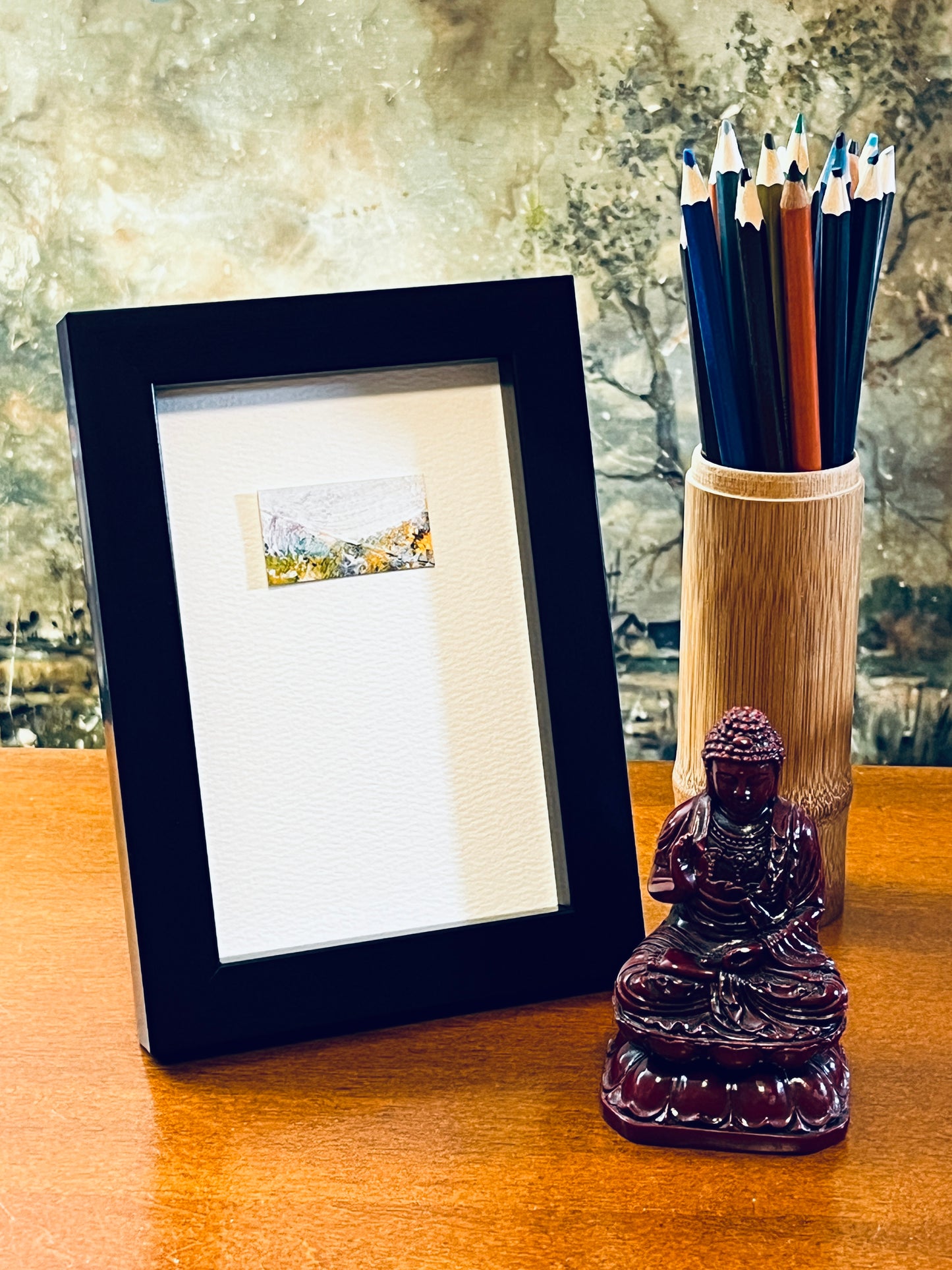 Original miniature abstract landscape painting featuring a mountain scene in spring foliage with a snowy backdrop. White, turquoise, yellow ochre, mint green orange and a touch of red is used. Acrylic paint on textured cold press watercolor paper, mounted on 4x6-inch watercolor paper in portrait orientation and displayed in a black frame next to a bamboo pencil holder and a small Buddha statue.