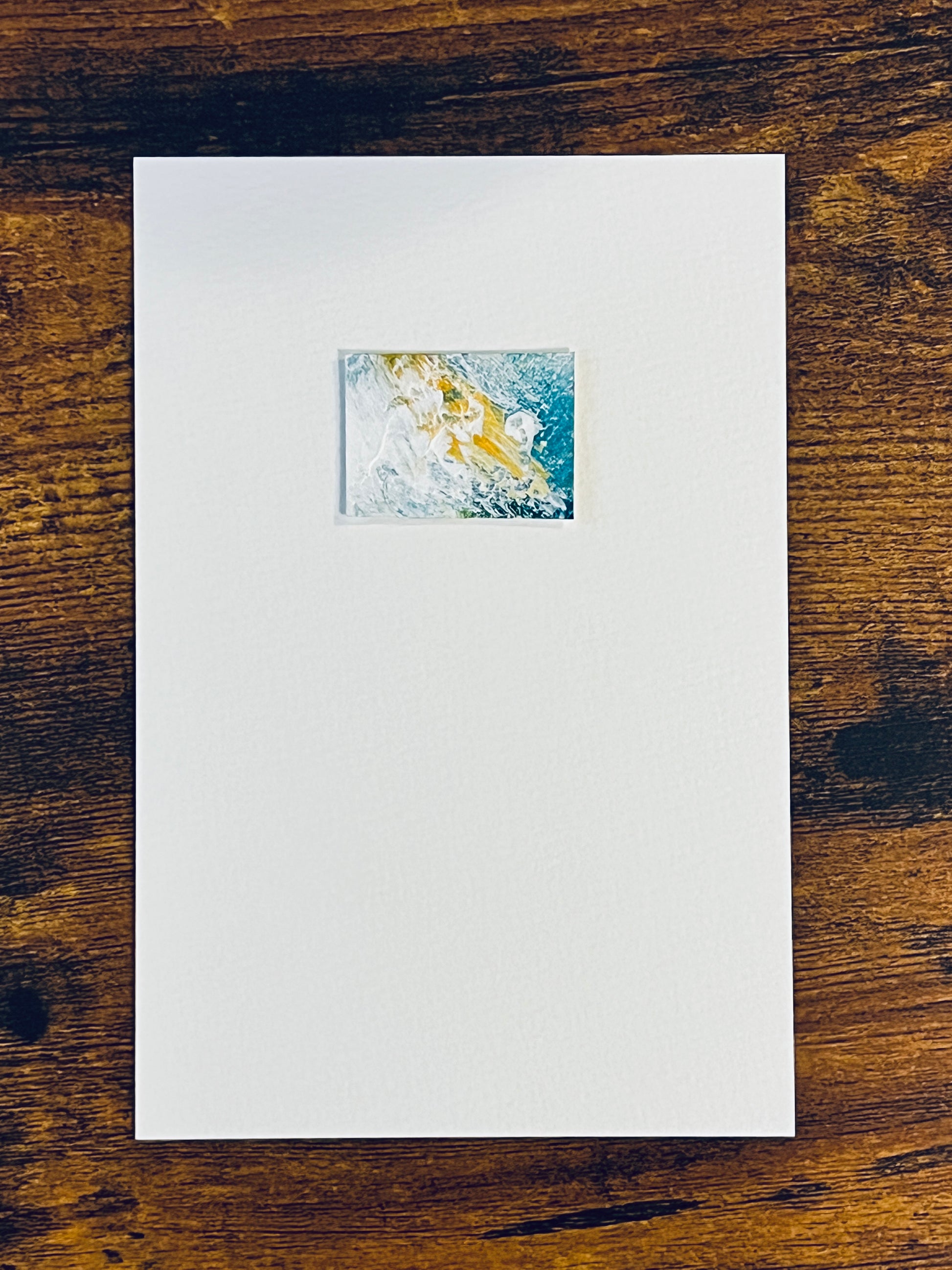 Original miniature abstract painting featuring yellow ochre teal green and white colors. Acrylic paint on textured cold press watercolor paper, mounted on 4x6-inch watercolor paper in portrait orientation and photographed on a wooden table.
