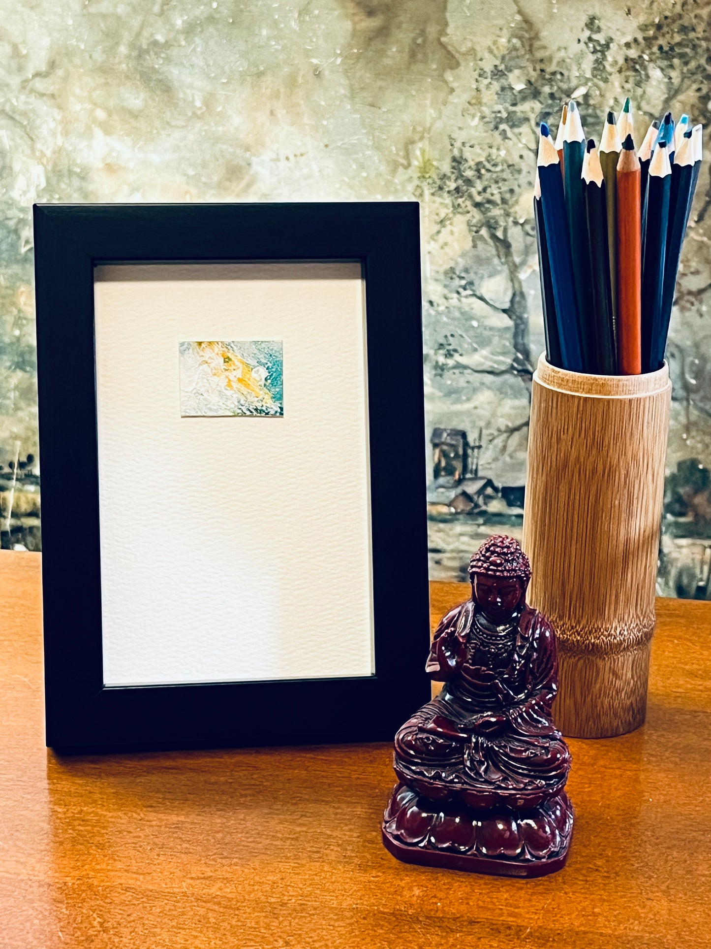 Original miniature abstract painting featuring yellow ochre teal green and white colors. Acrylic paint on textured cold press watercolor paper, mounted on 4x6-inch watercolor paper in portrait orientation and displayed in a black frame next to a bamboo pencil holder and a small Buddha statue.