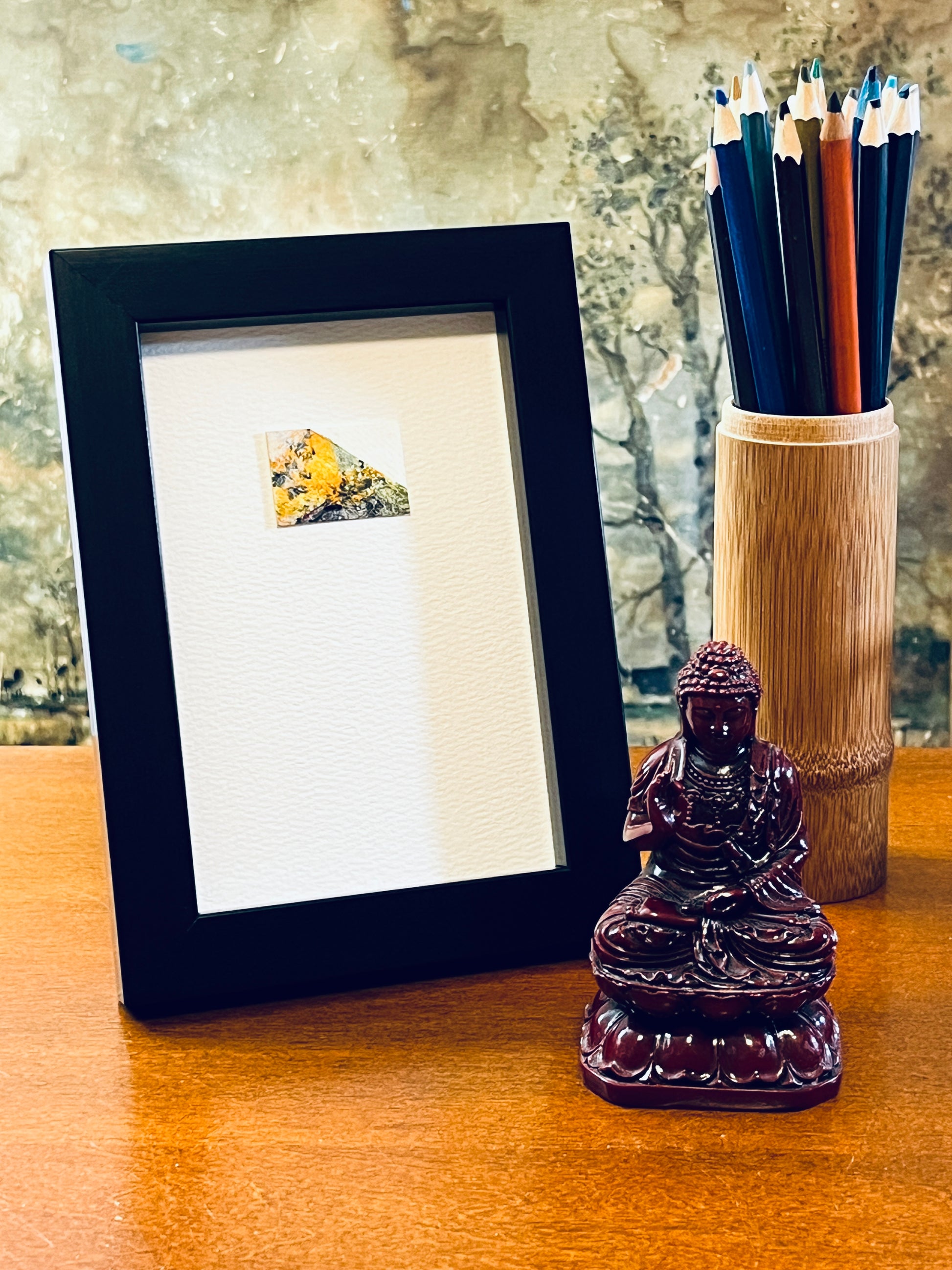 Original miniature abstract painting featuring yellow ochre, mint green grey and a touch of red paint are used. Acrylic paint on textured cold press watercolor paper, mounted on 4x6-inch watercolor paper in portrait orientation and displayed in a black frame next to a bamboo pencil holder and a small Buddha statue.