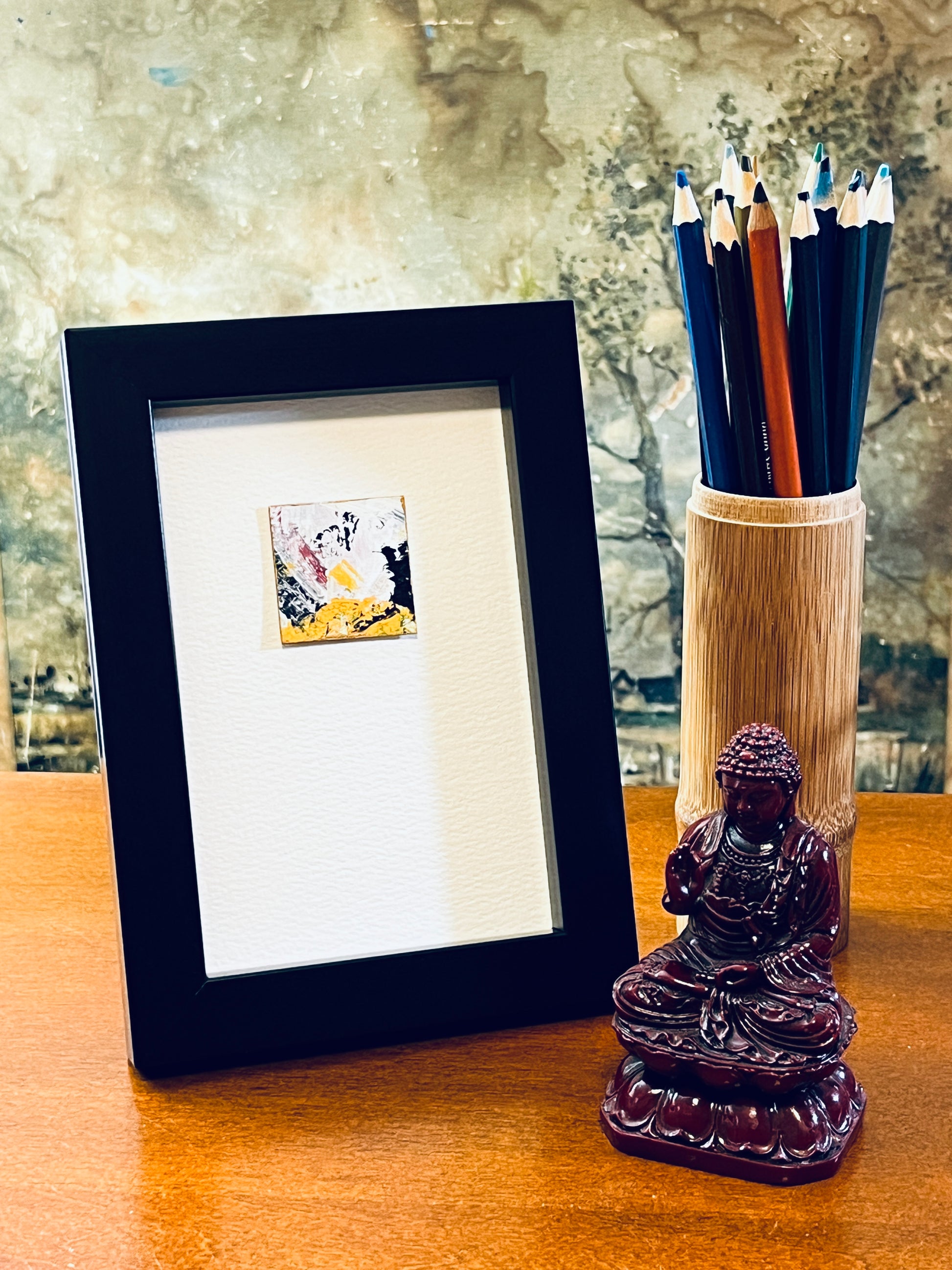 Original miniature abstract painting featuring white, yellow ochre, black, red and a touch of green paint. Acrylic paint with gold wax metallic edges on textured cold press watercolor paper, mounted on 4x6-inch watercolor paper in portrait orientation and displayed in a black frame next to a bamboo pencil holder and a small Buddha statue.