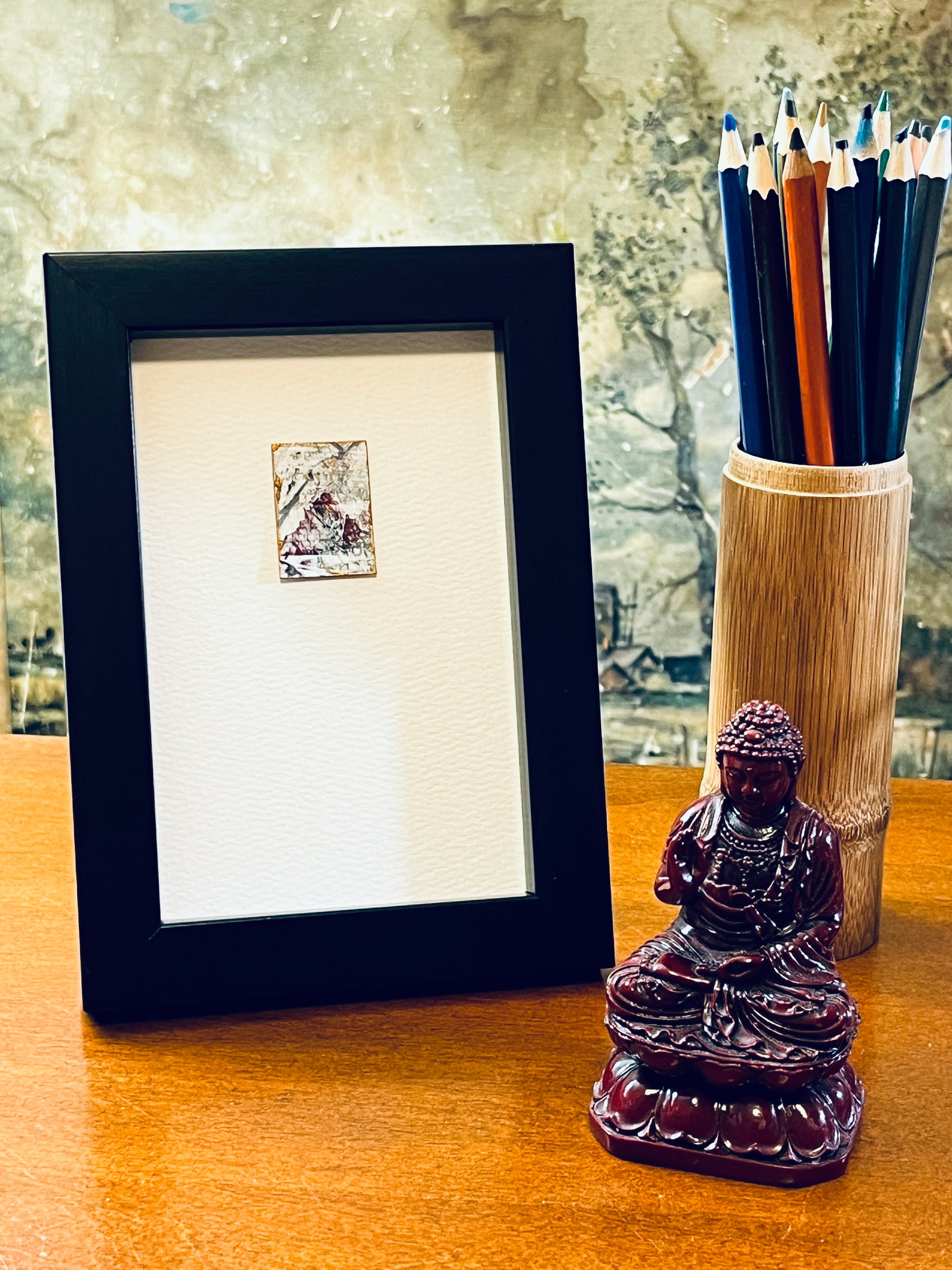 Original miniature abstract painting featuring white, grey and red acrylic paint on textured cold press watercolor paper, displayed in a black 4x6-inch frame photographed next to a bamboo pencil holder and a small Buddha statue.