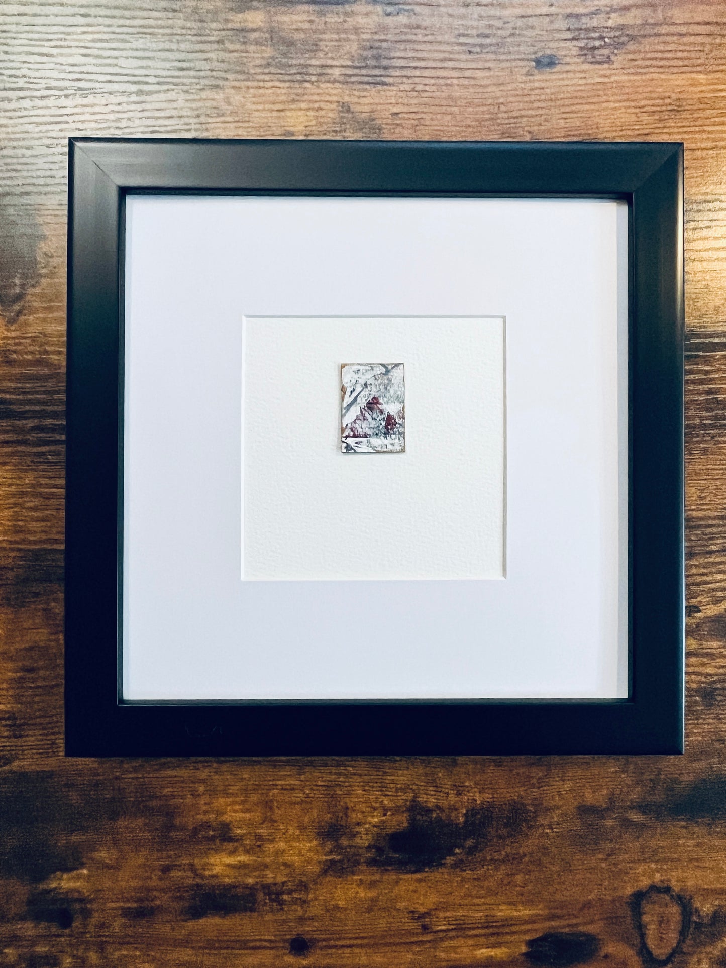 Original miniature abstract painting featuring white, grey and red acrylic paint on textured cold press watercolor paper, displayed in a black 8x8-inch frame with mat, photographed on a wooden table.