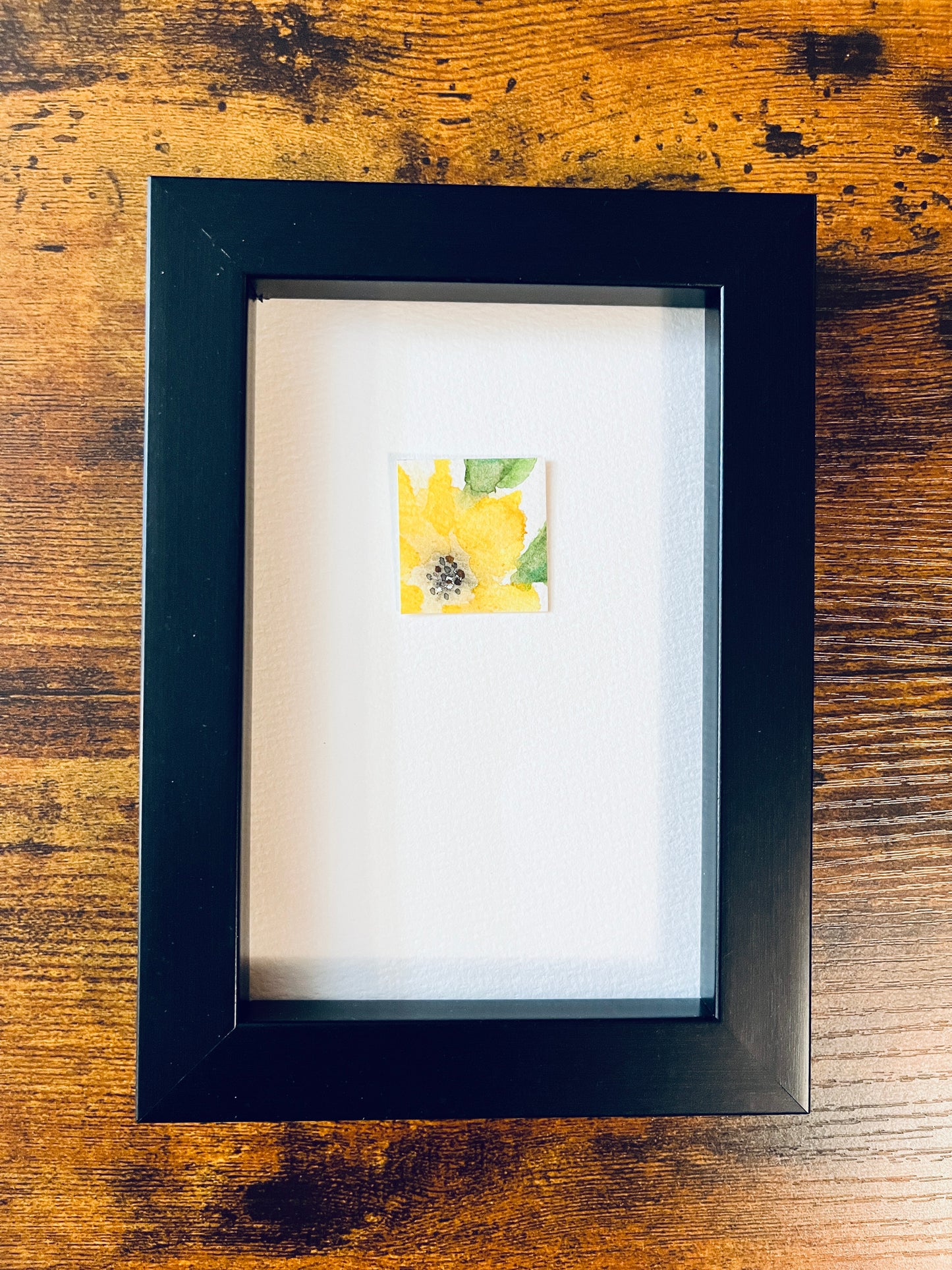 Original artwork by Anet Skillin. This miniature abstract painting features a yellow thunbergia flower and green leaves. Painted on textured cold-press watercolor paper; mounted on 4x6-inch paper and photographed in a black 4x6-inch frame against a wooden table.