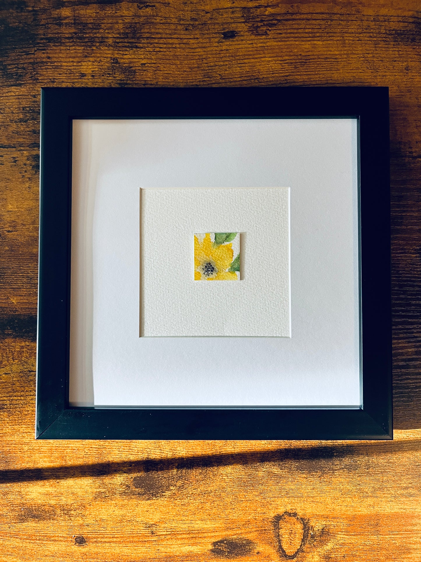 Original artwork by Anet Skillin. This miniature abstract painting features a yellow thunbergia flower and green leaves. Painted on textured cold-press watercolor paper; mounted on 4x6-inch paper and photographed in a black 8x8-inch frame against a wooden table.