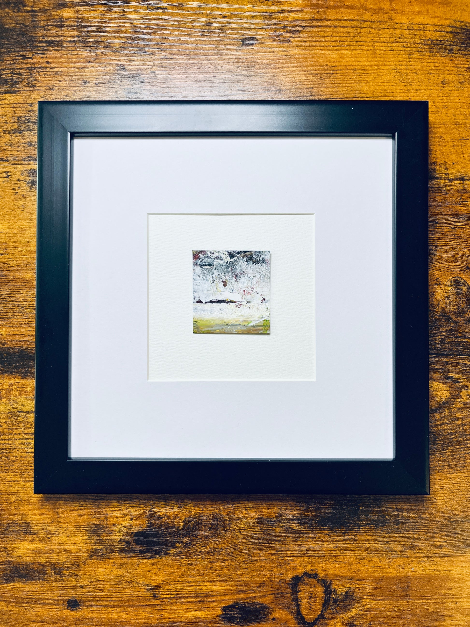 Original artwork by Anet Skillin. This miniature abstract landscape painting is painted on recycled packaging in white, red, yellow ochre and green colors; mounted on 4x6-inch paper and photographed in a black 8x8-inch frame against a wooden table.