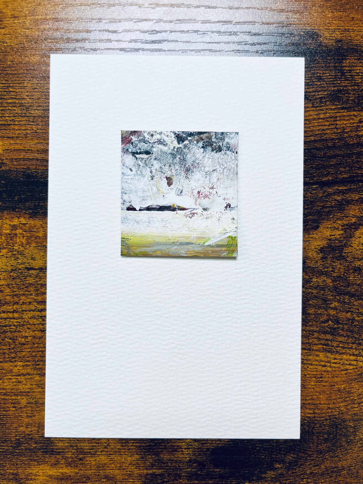 Original artwork by Anet Skillin. This miniature abstract landscape painting is painted on recycled packaging in white, red, yellow ochre and green colors; mounted on 4x6-inch paper and photographed against a wooden table.